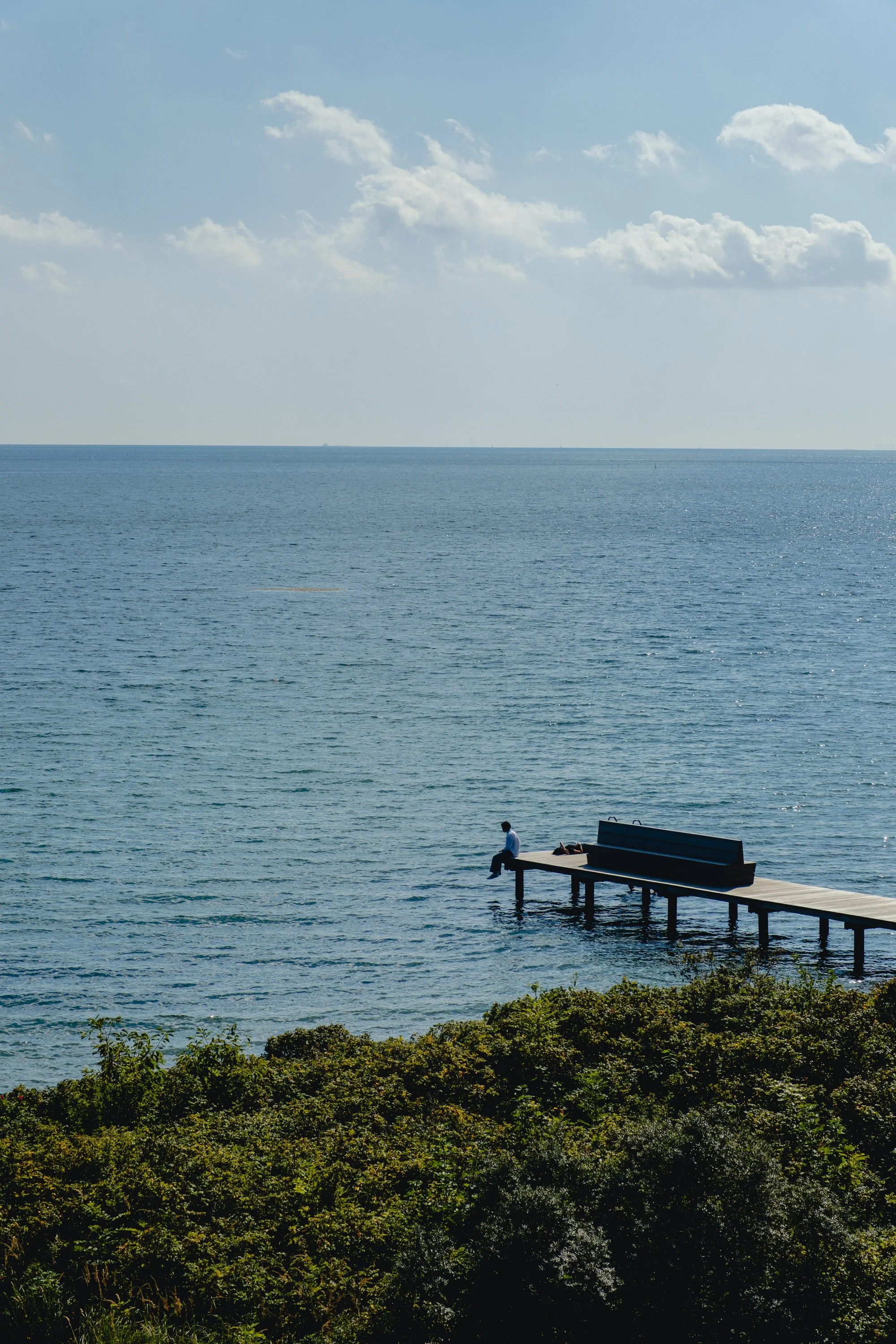 A person sitting on the edge of a pier or dock extending over a body of water, with a view of the ocean, sky, and clouds.