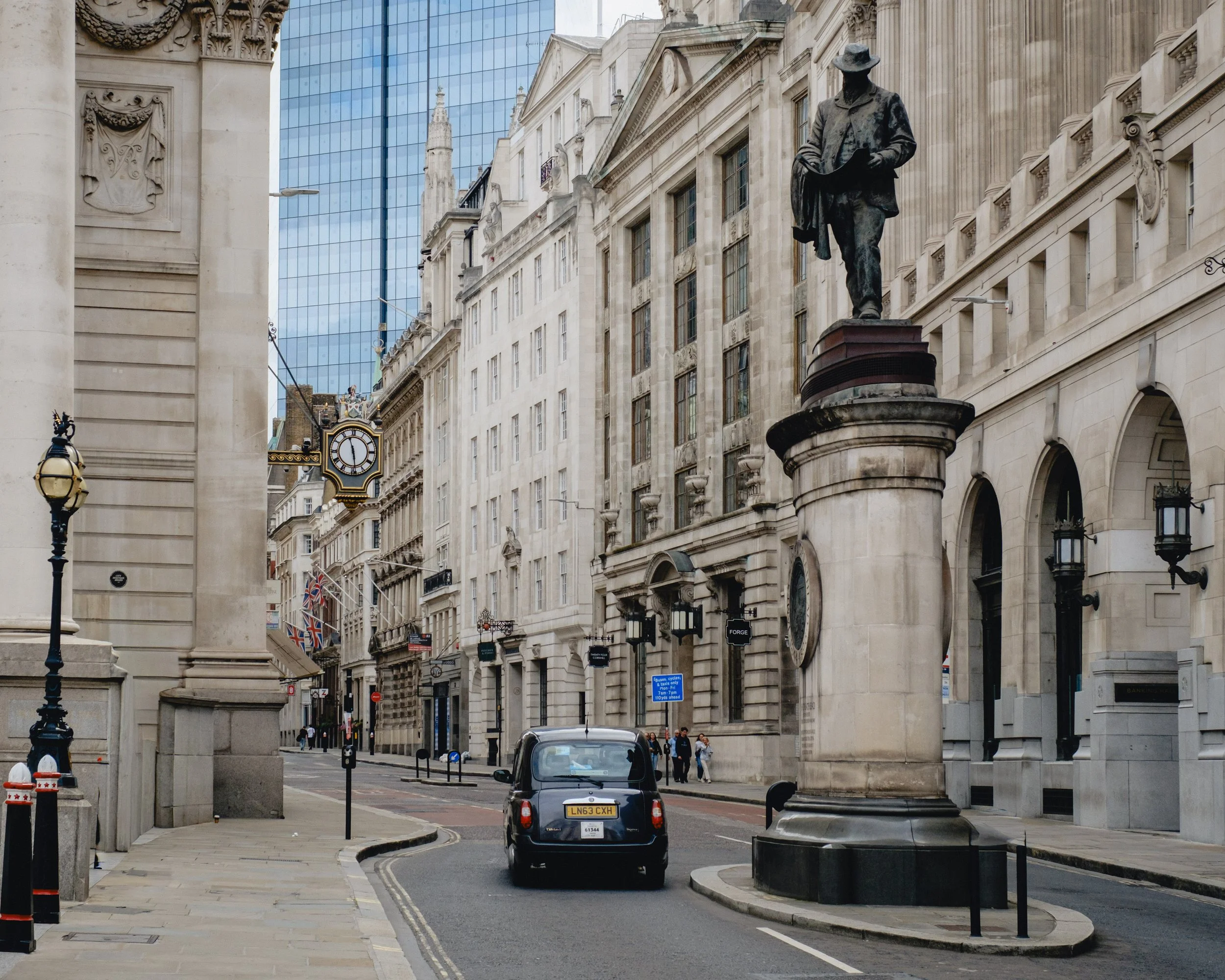 A city street with historic and modern buildings, a statue on a tall pedestal, a black taxi cab, a clock showing 2 o'clock, and a few pedestrians.
