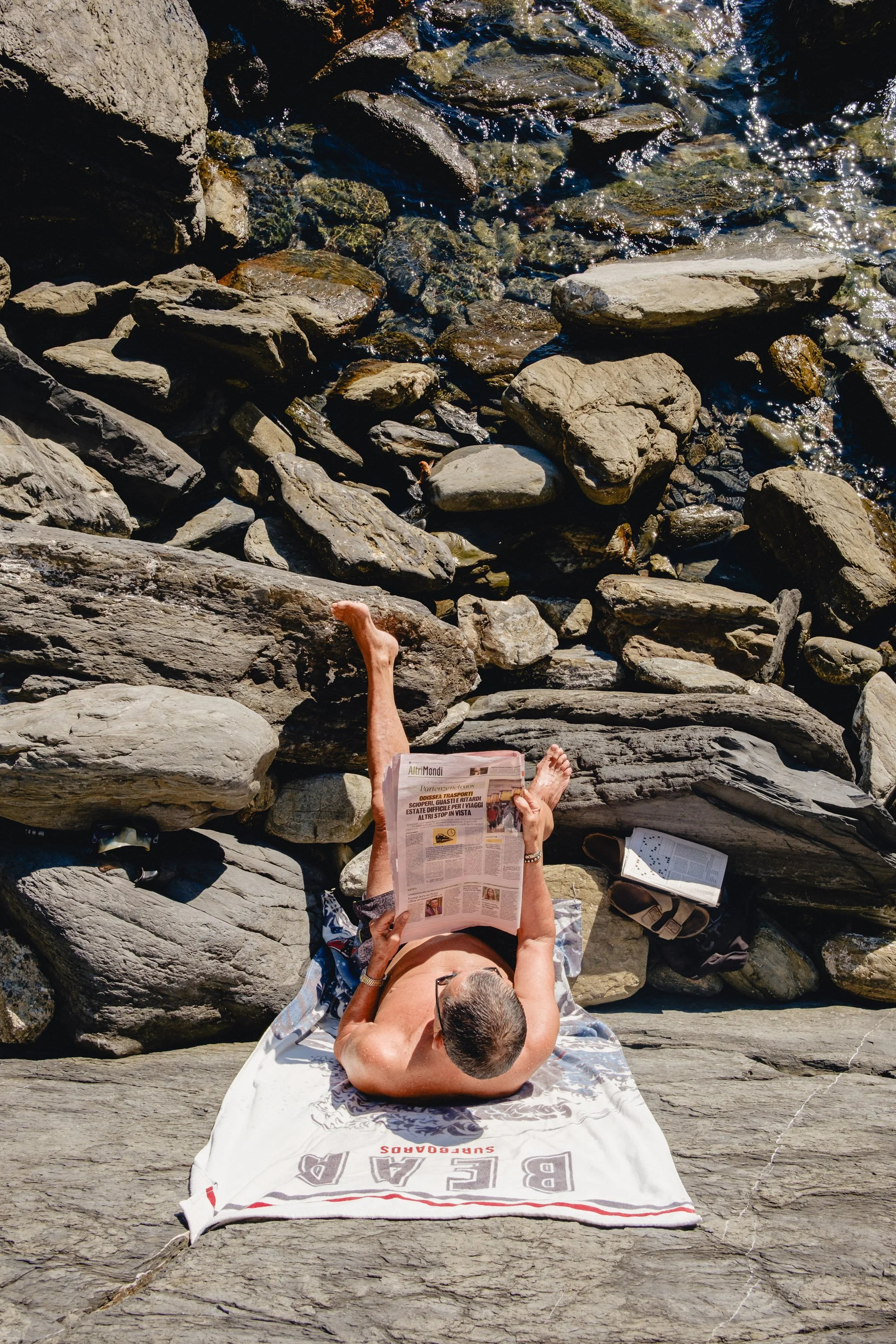 A person lying on a towel on a large flat rock by a rocky shoreline, reading a newspaper during the day.
