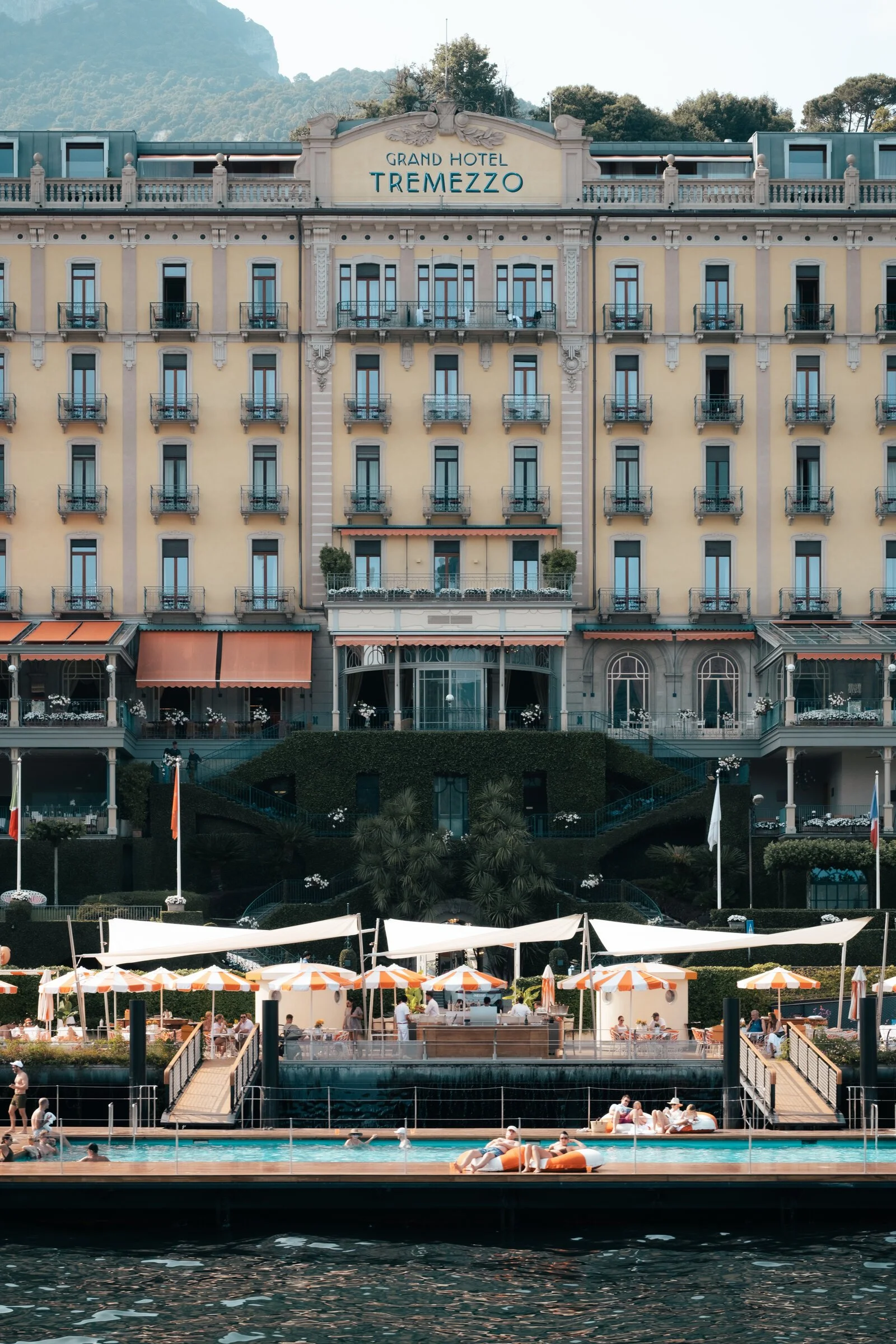 The pale yellow facade of the Grand Hotel Tremezzo rises above its floating swimming pool and orange umbrellas on Lake Como