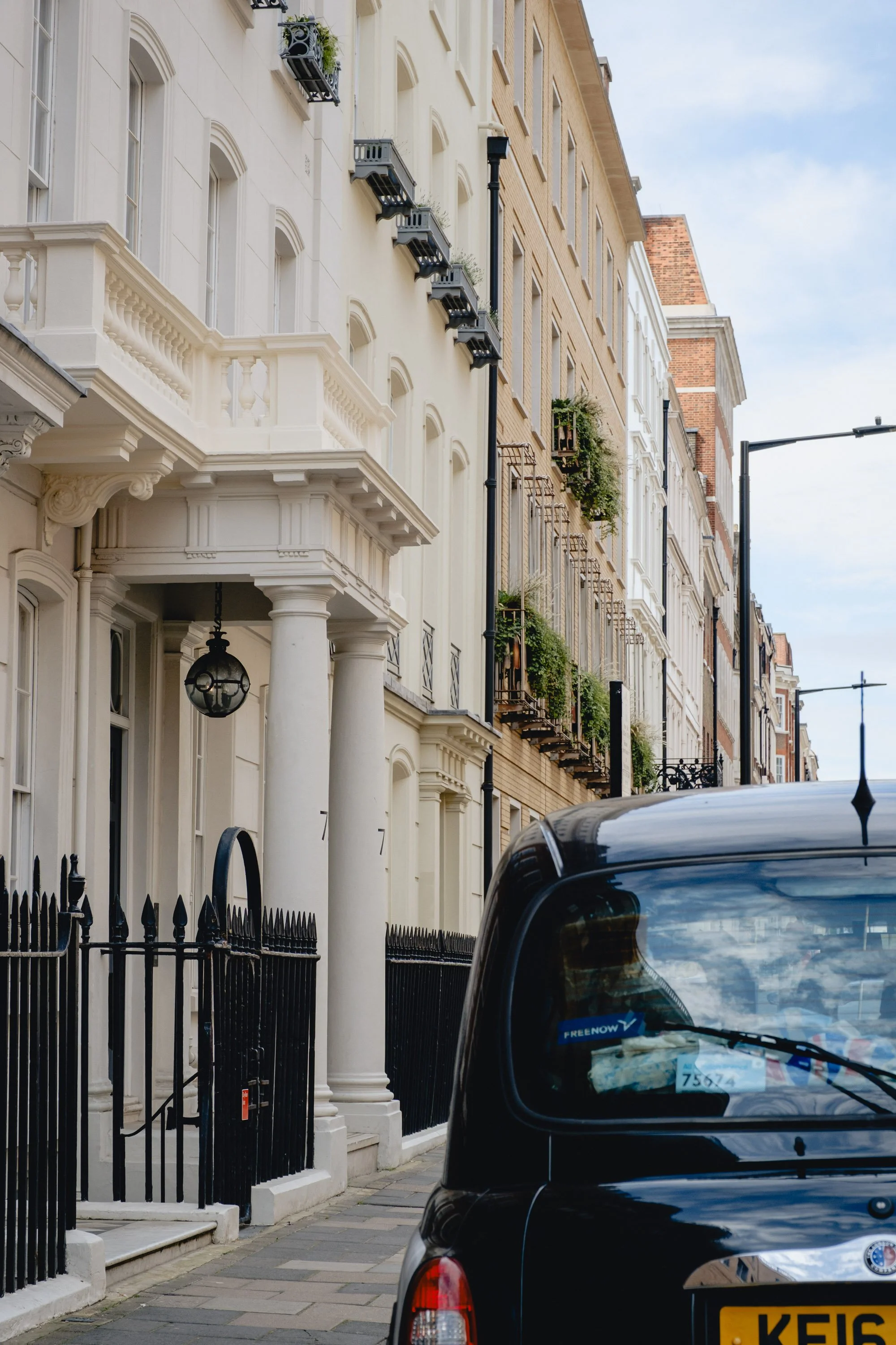 Street view showing row of historic townhouses with ornate facades, balconies with potted plants, and iron gates, with a black cab parked on the street.