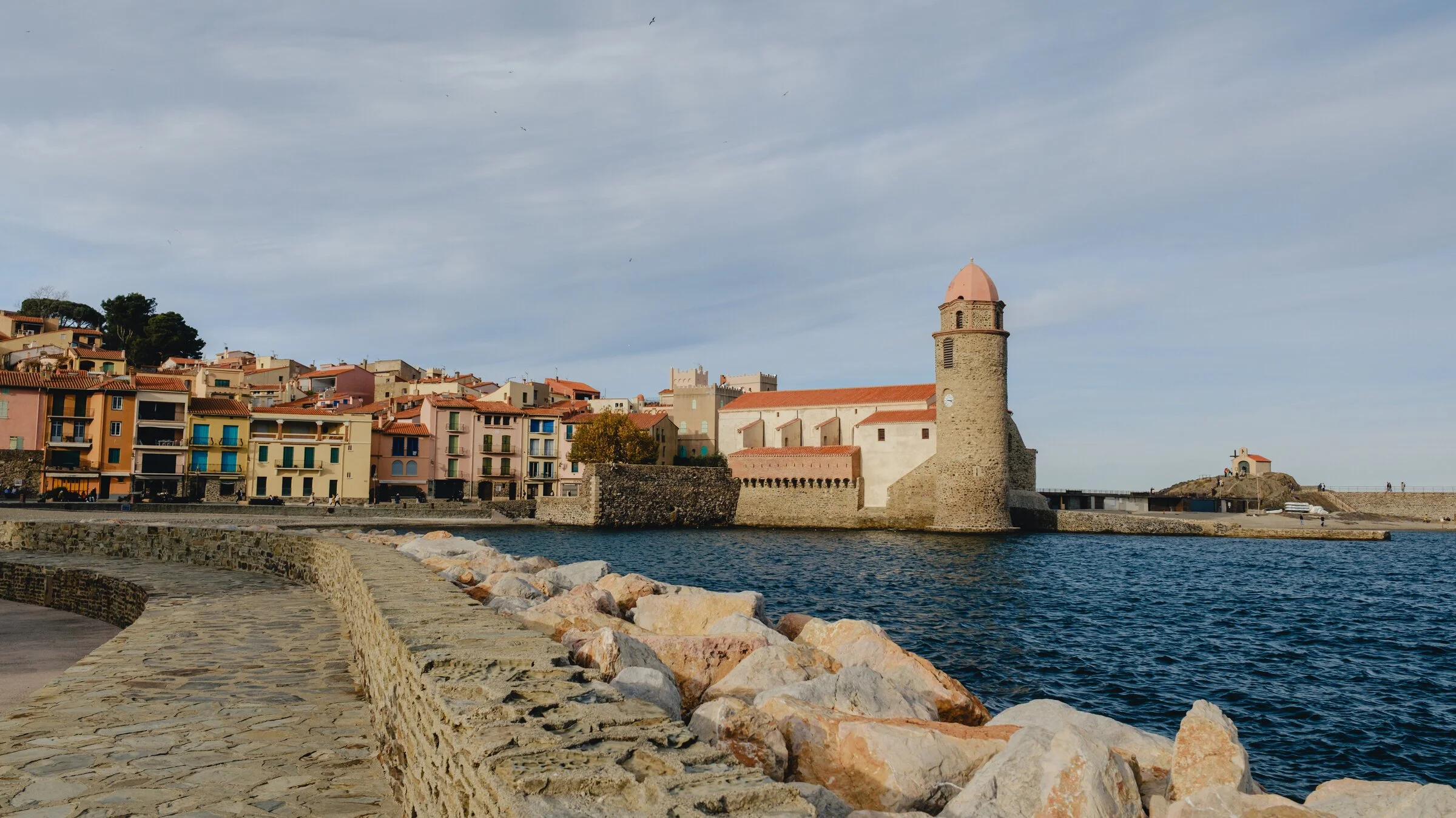 Collioure's pastel harborfront buildings and church tower seen across the water from a stone jetty