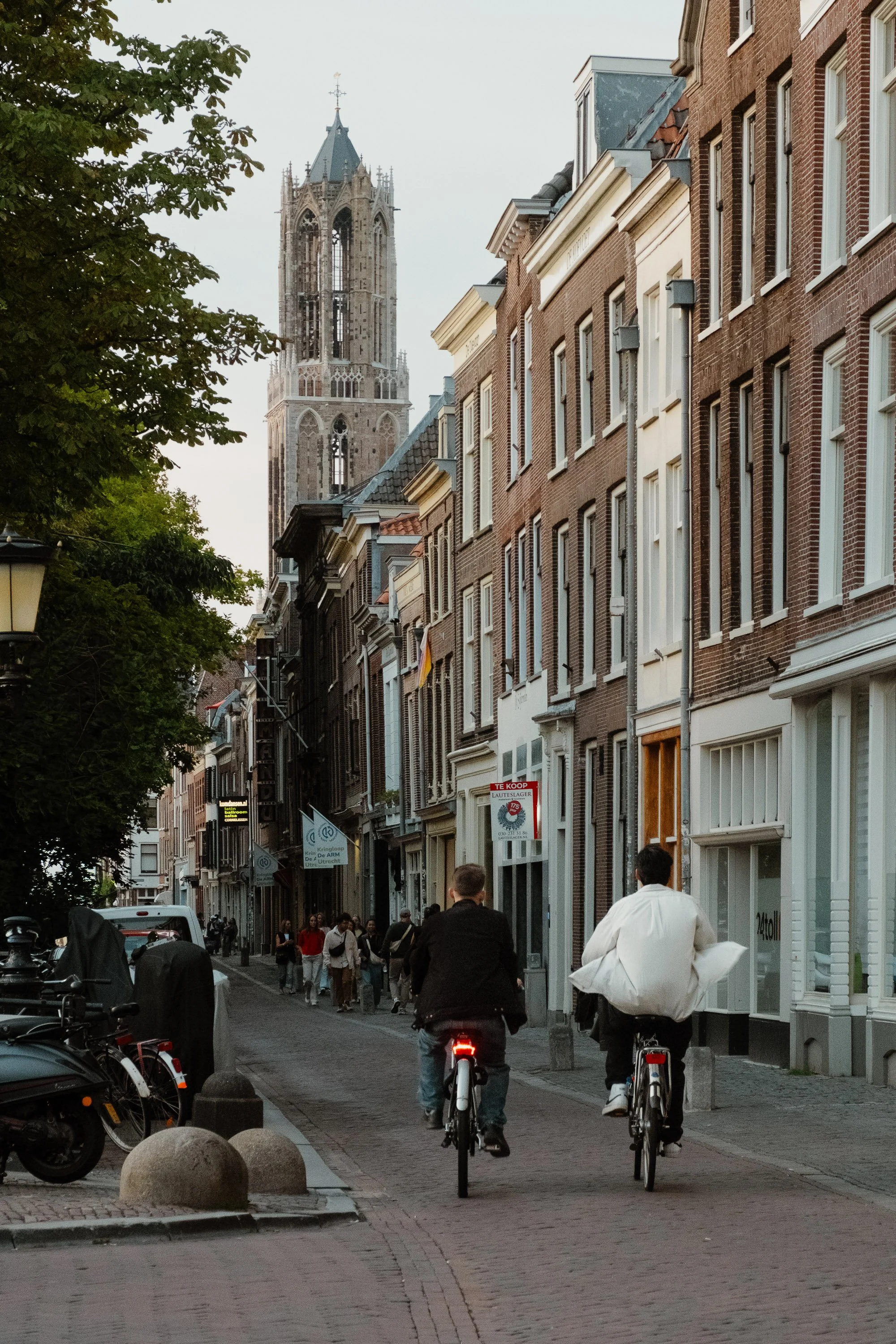 A street scene with people riding bicycles and walking along a narrow cobblestone street lined with historic brick buildings. There is a tall church tower in the background and some trees on the left side of the street.