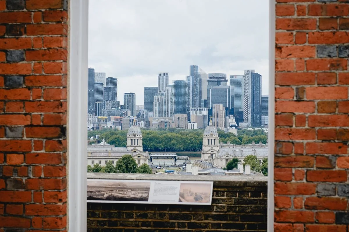 Canary Wharf's towers seen through a gap in a brick wall, Greenwich domes and park below