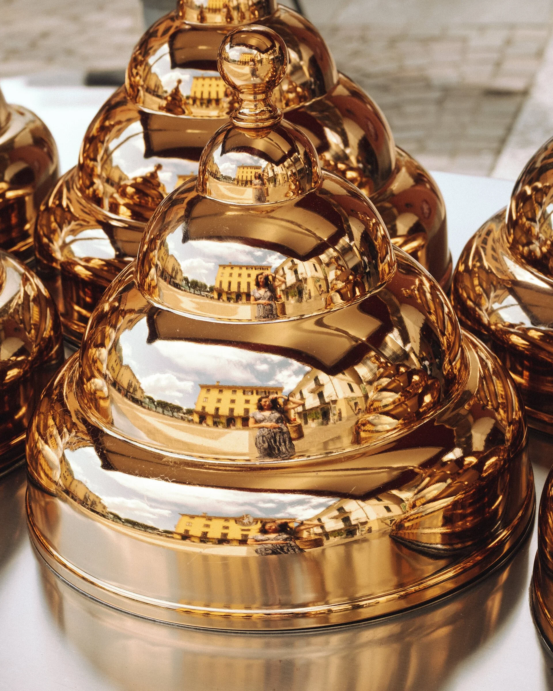 A row of polished gold dome cloches reflect a distorted, warped image of a yellow building and passersby