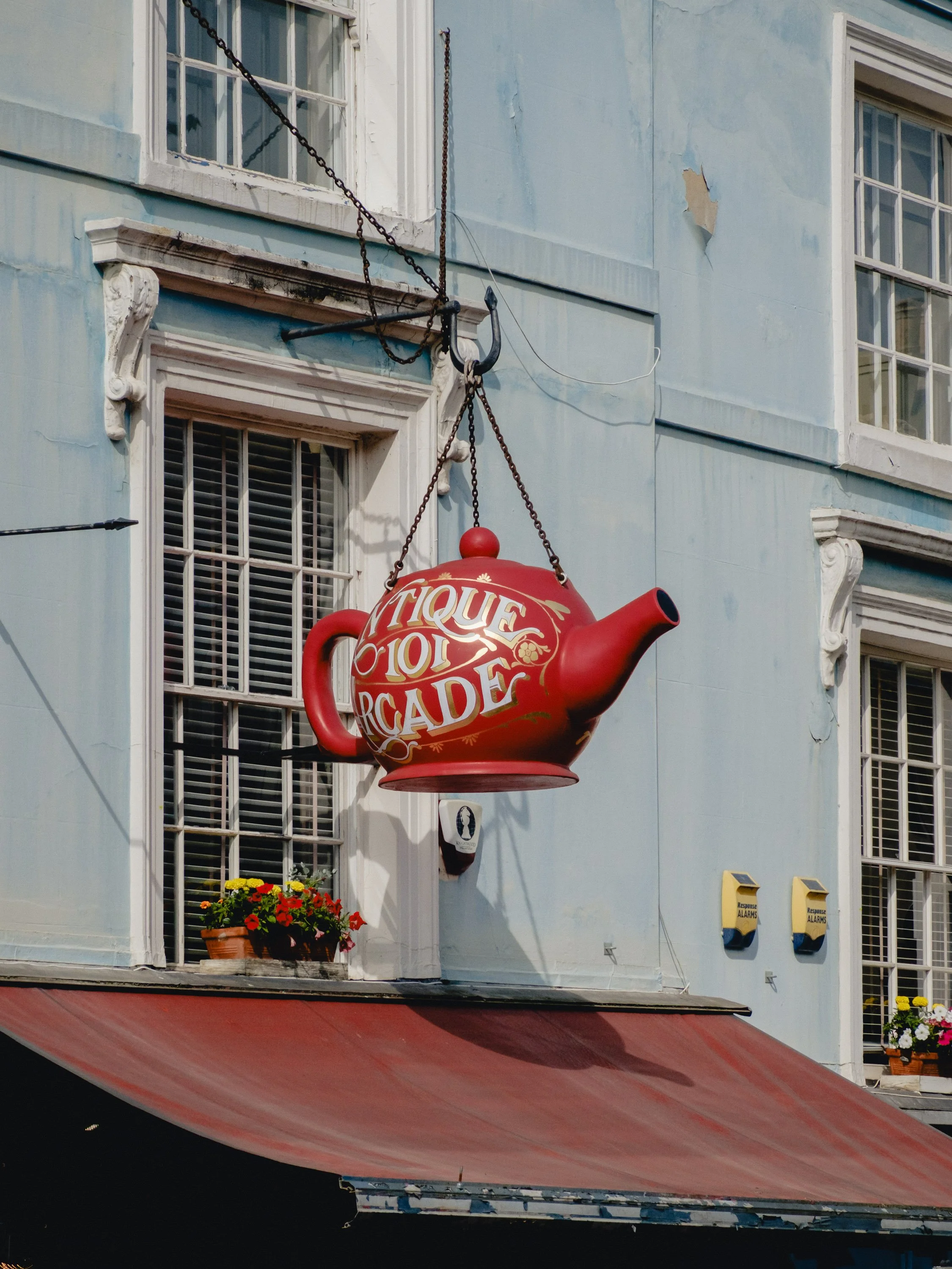 A floating red teapot-shaped sign with gold and white writing says 'Tique Locale Arcade,' hanging from chains on a light blue building with white trim. Below is a window with a flowerbox containing red and yellow flowers.