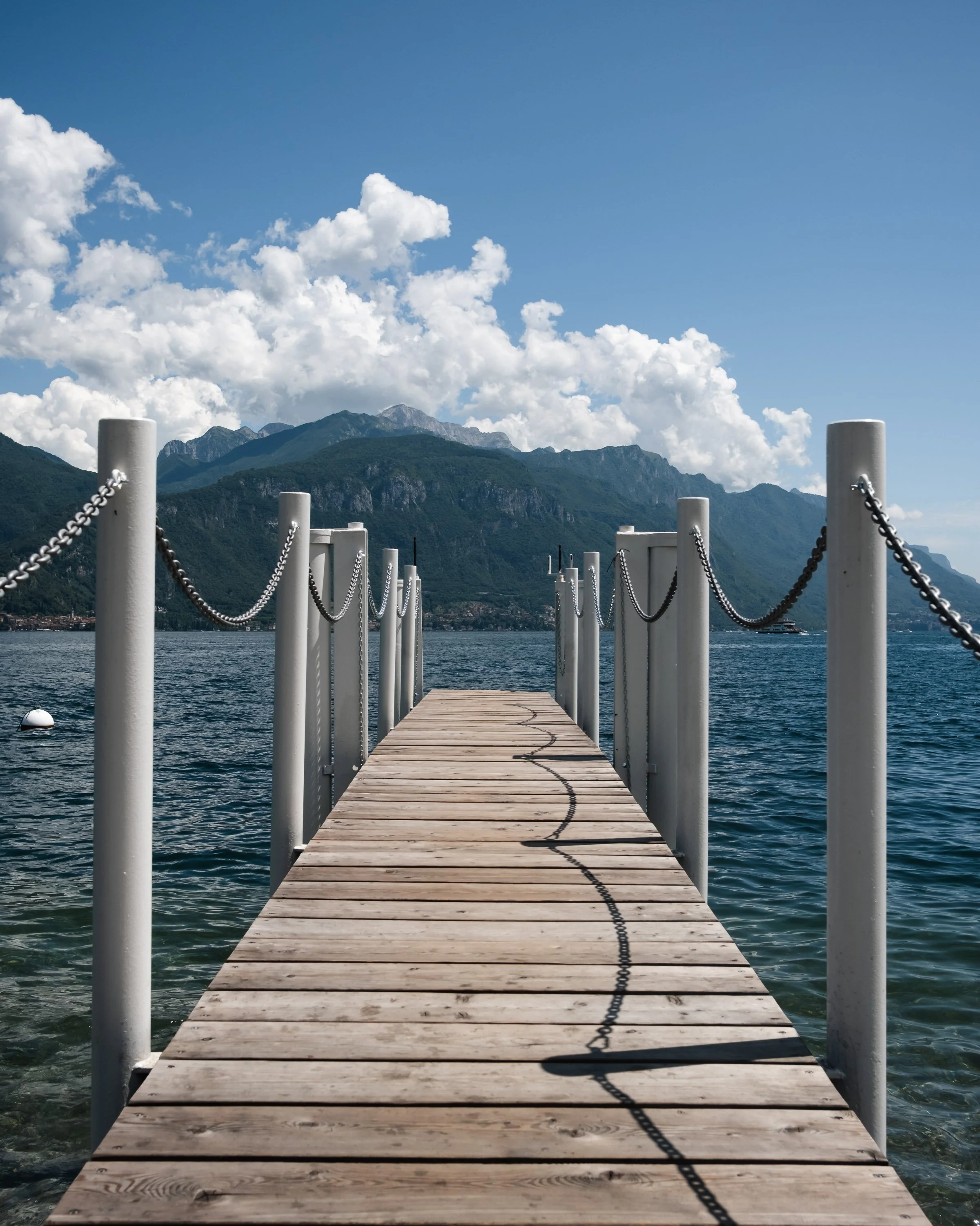 Wooden dock extending over water with chain railing, mountains in the background, blue sky, and clouds.