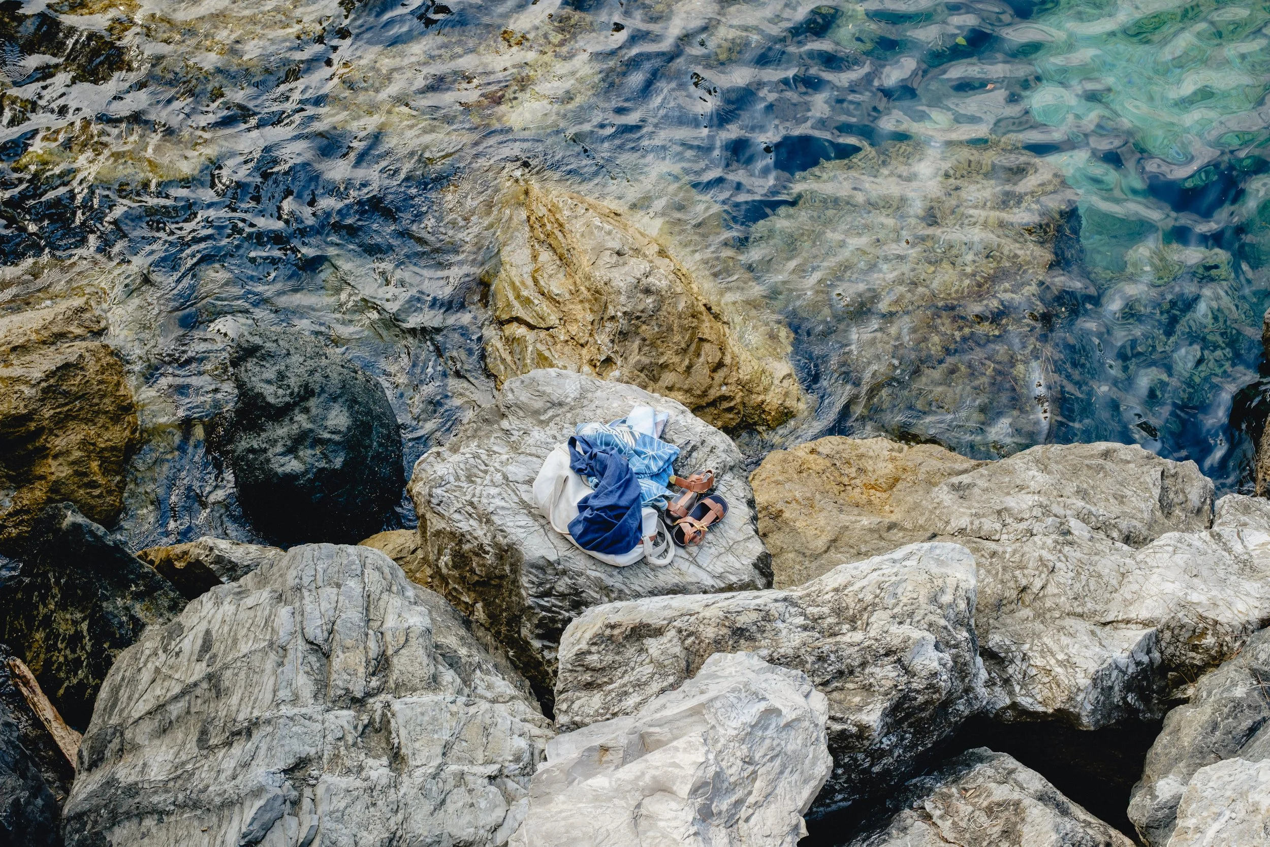Clothes and sandals on a large rock by a rocky shoreline with clear blue water.