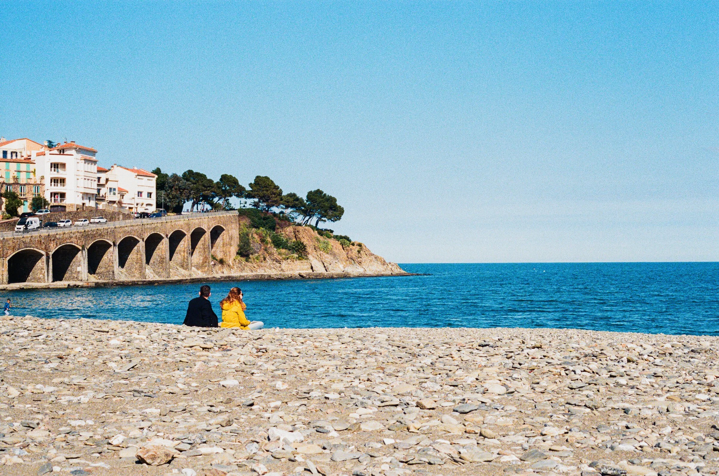 A couple sits on a pebble beach beneath the stone arches of a coastal railway viaduct on the Cote Vermeille