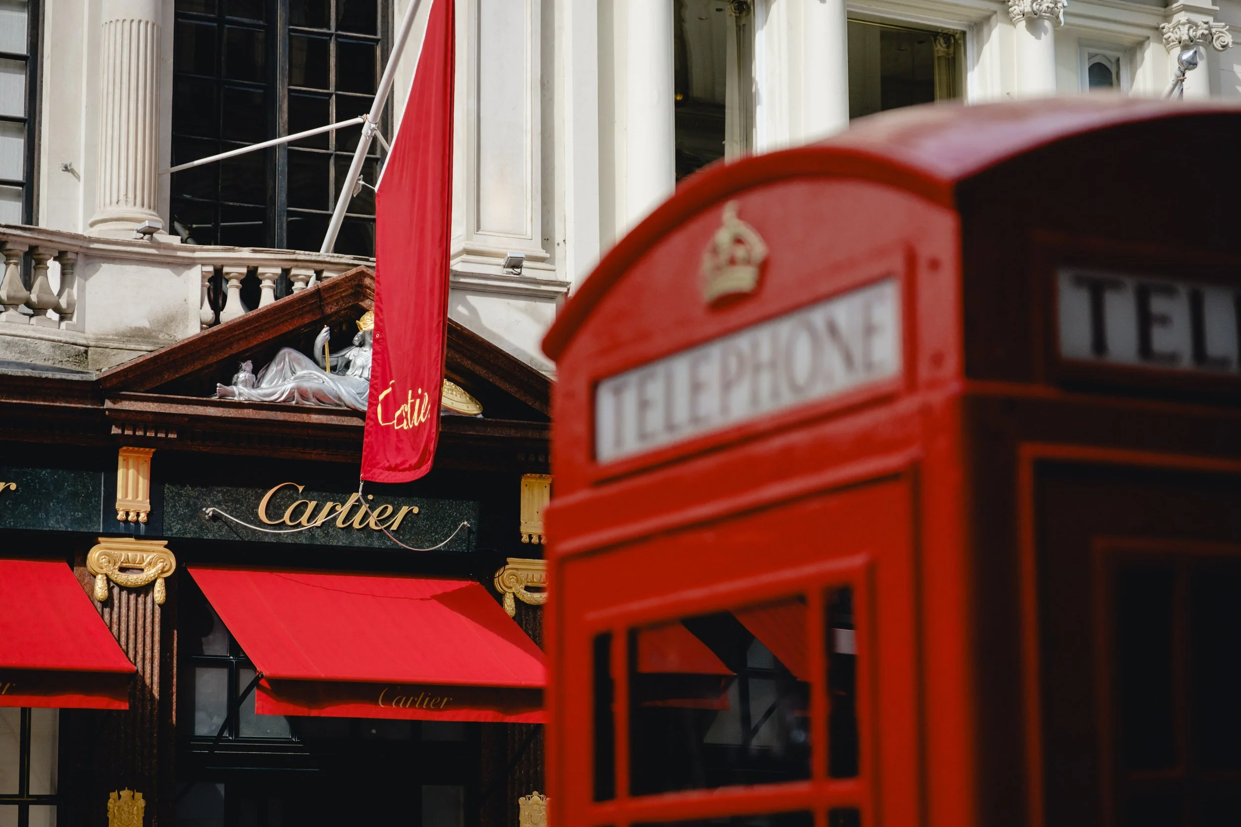 A red British telephone booth in front of an elegant storefront with the Cartier logo, red awnings, and a red flag with gold text.