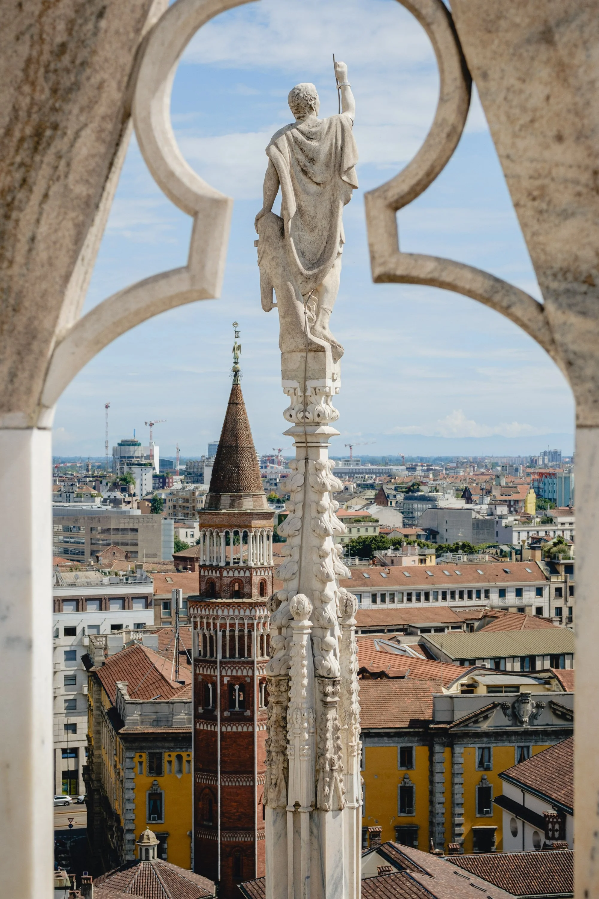 Statue of a robed figure holding a spear, seen from behind, on a tall decorative pillar, overlooking a cityscape with historic buildings and a blue sky.
