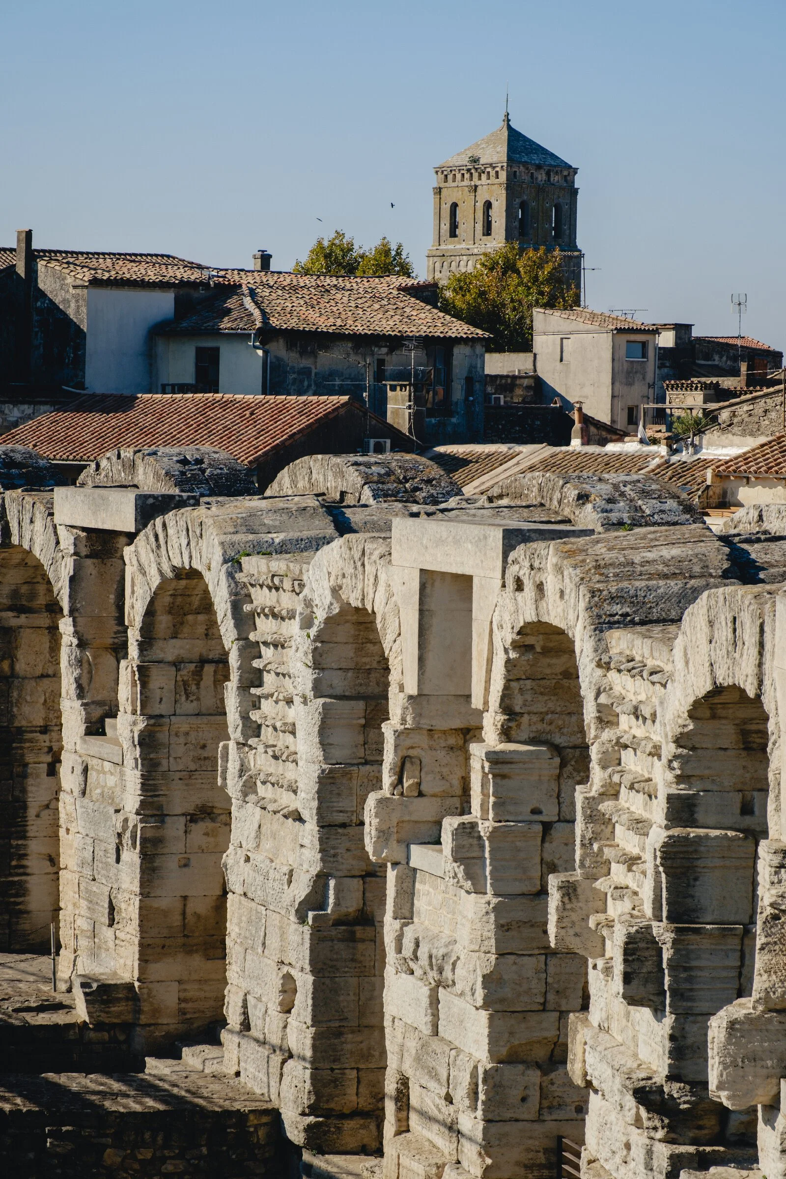 Weathered stone arches of the Roman amphitheatre in Arles rise above the town's rooftops and a bell tower