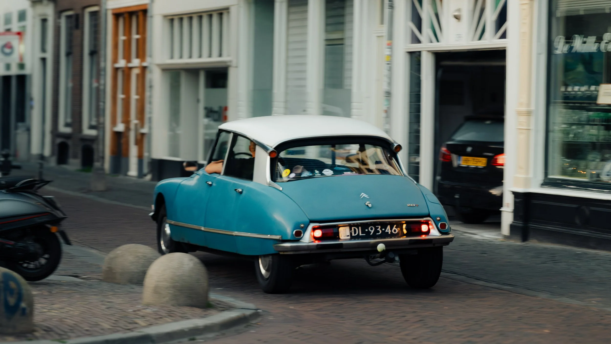 A vintage blue and white Citroën 2CV car driving on a city street with shops and parked cars.