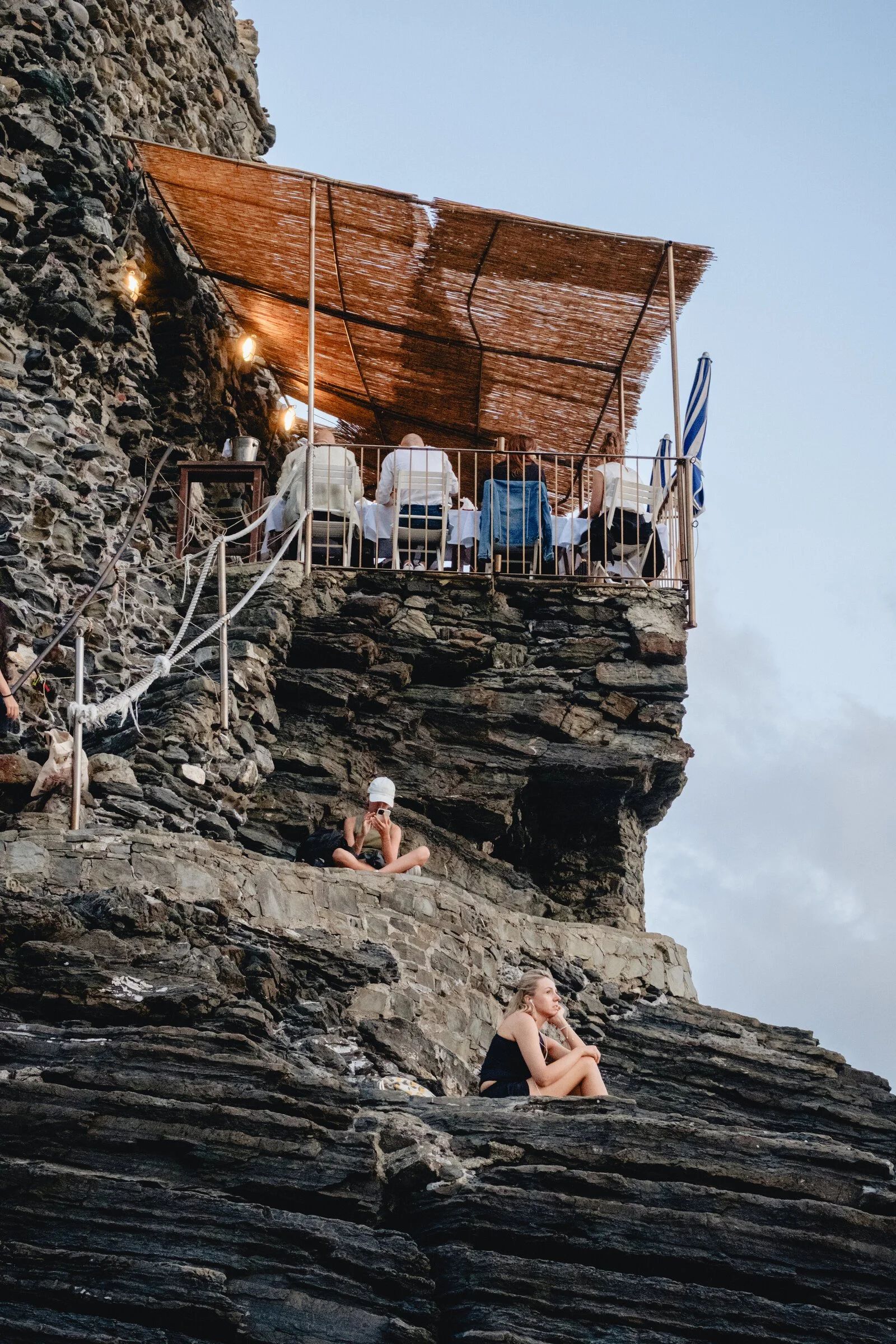 Diners on a cliffside terrace at dusk above Vernazza, two women sitting on the rocks below