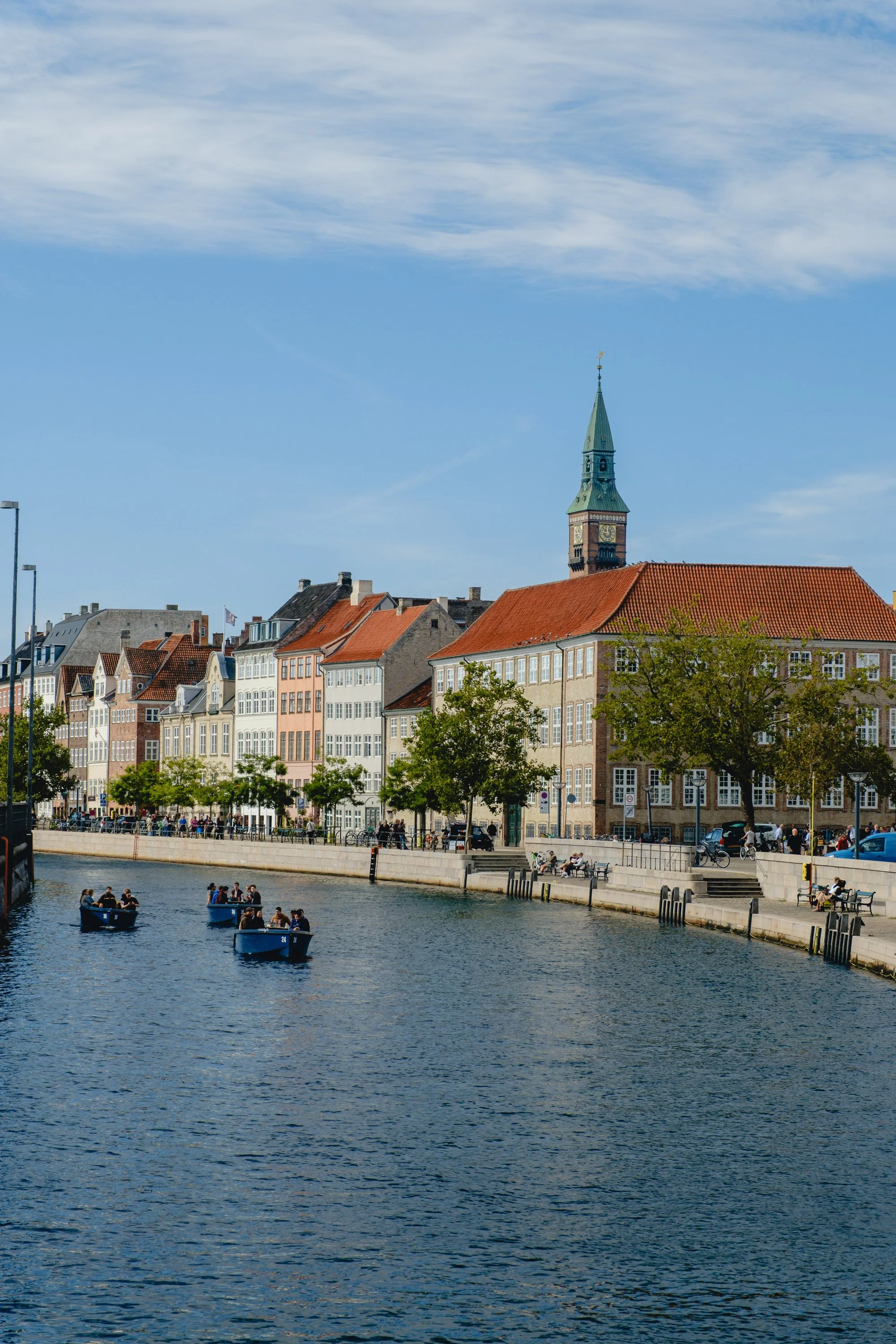 View of colorful European buildings along a river with boats, trees, and a clock tower under a partly cloudy sky.