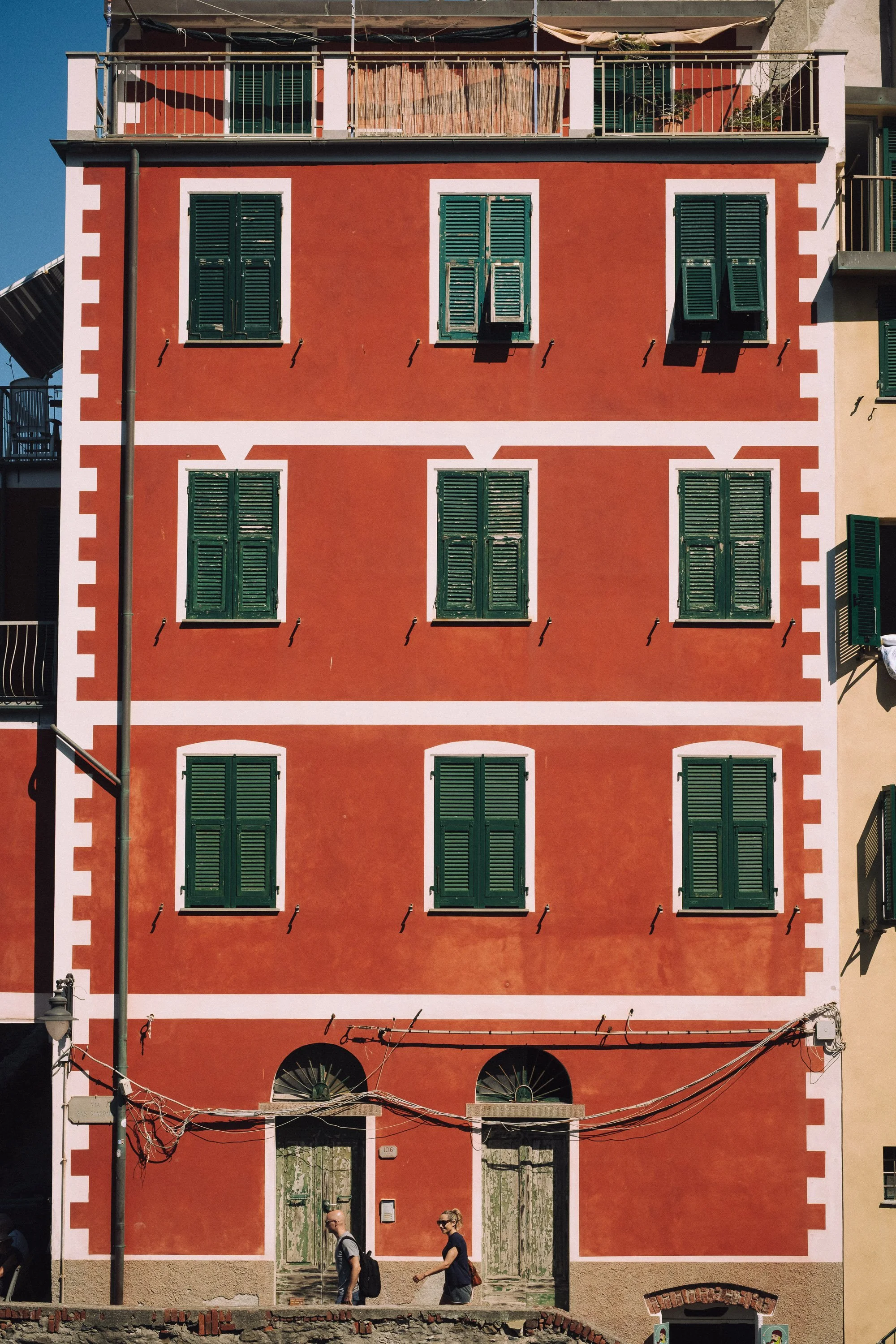 Red building with green shutters on multiple windows, some partially open, and white trim, with two people walking in front.