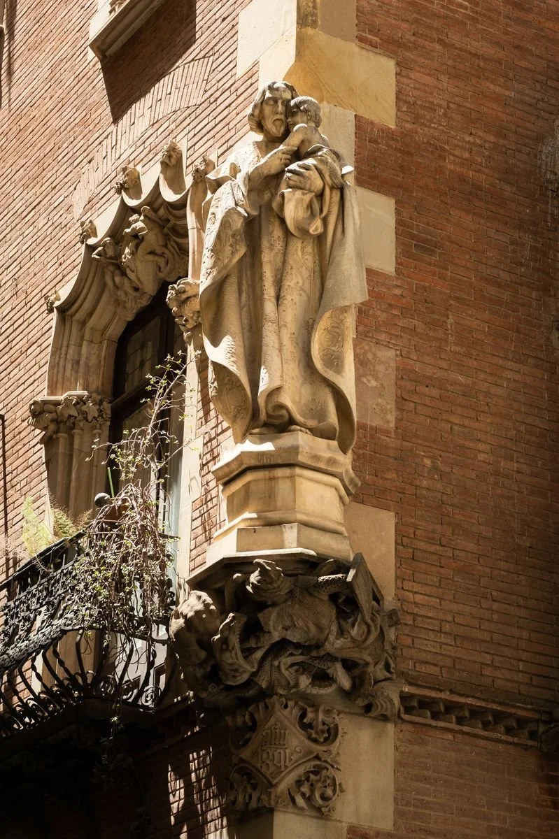 A carved stone saint stands on a brick building's corner, framed by a Gothic window