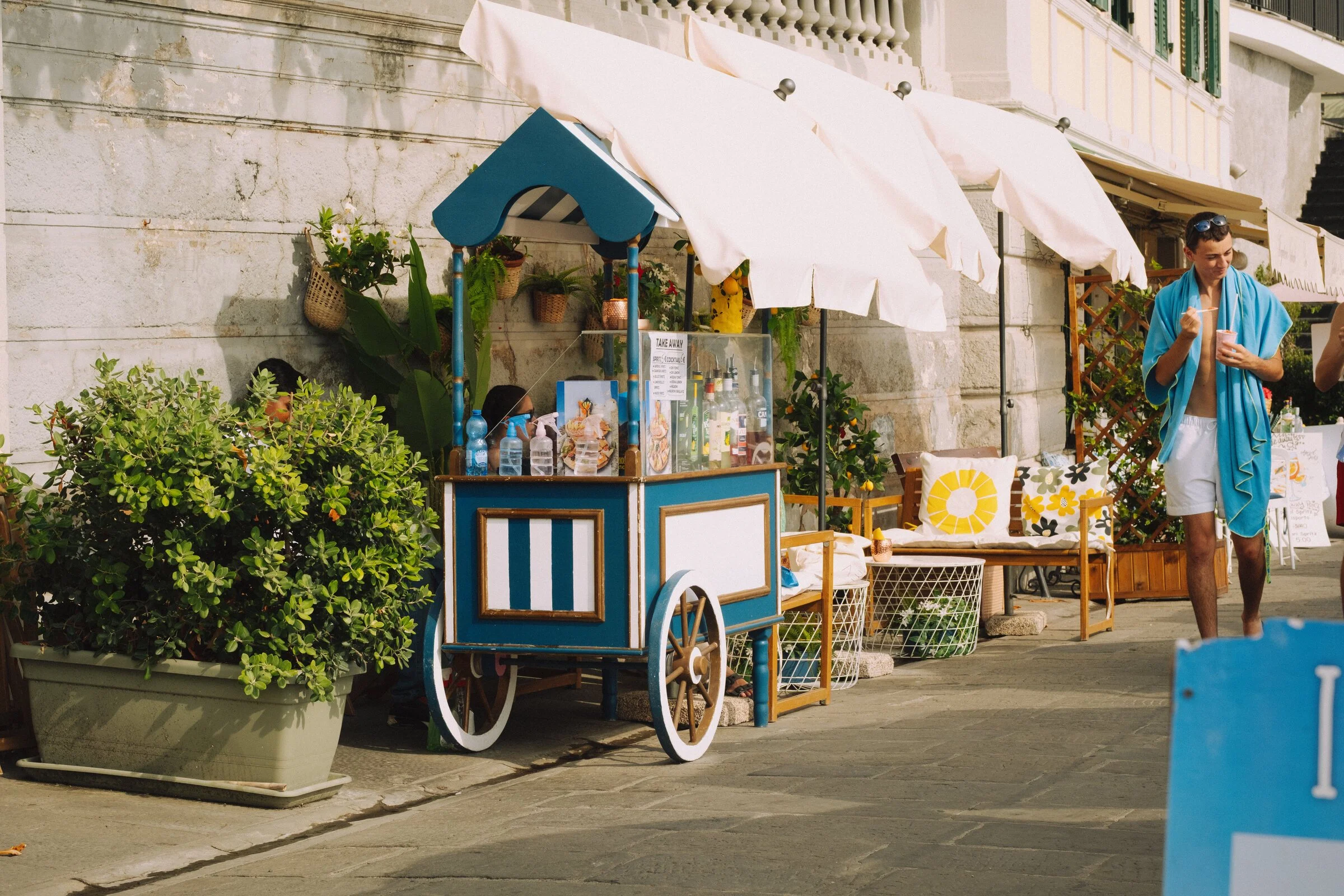 A blue vintage vending cart with an awning sits along a promenade beside a man in a towel