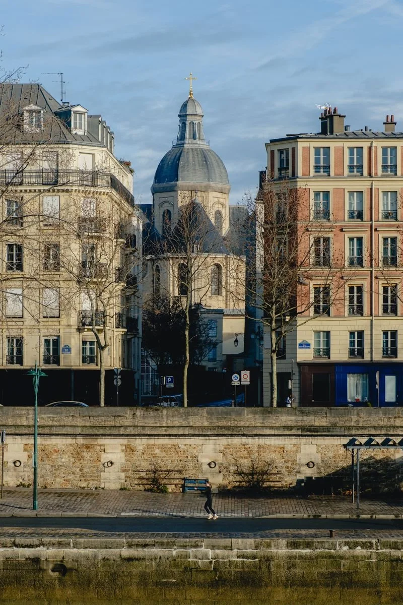 A domed church rises between apartment buildings above a quiet riverside quay where a jogger runs