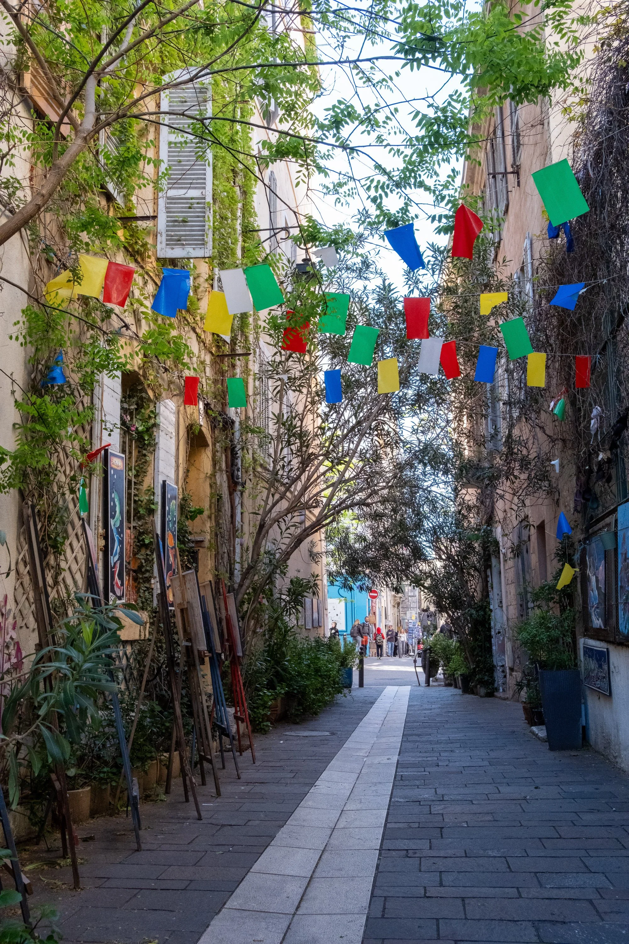A narrow pedestrian street decorated with colorful paper flags hanging overhead, with trees, potted plants, artwork, and people visible at the end of the street.