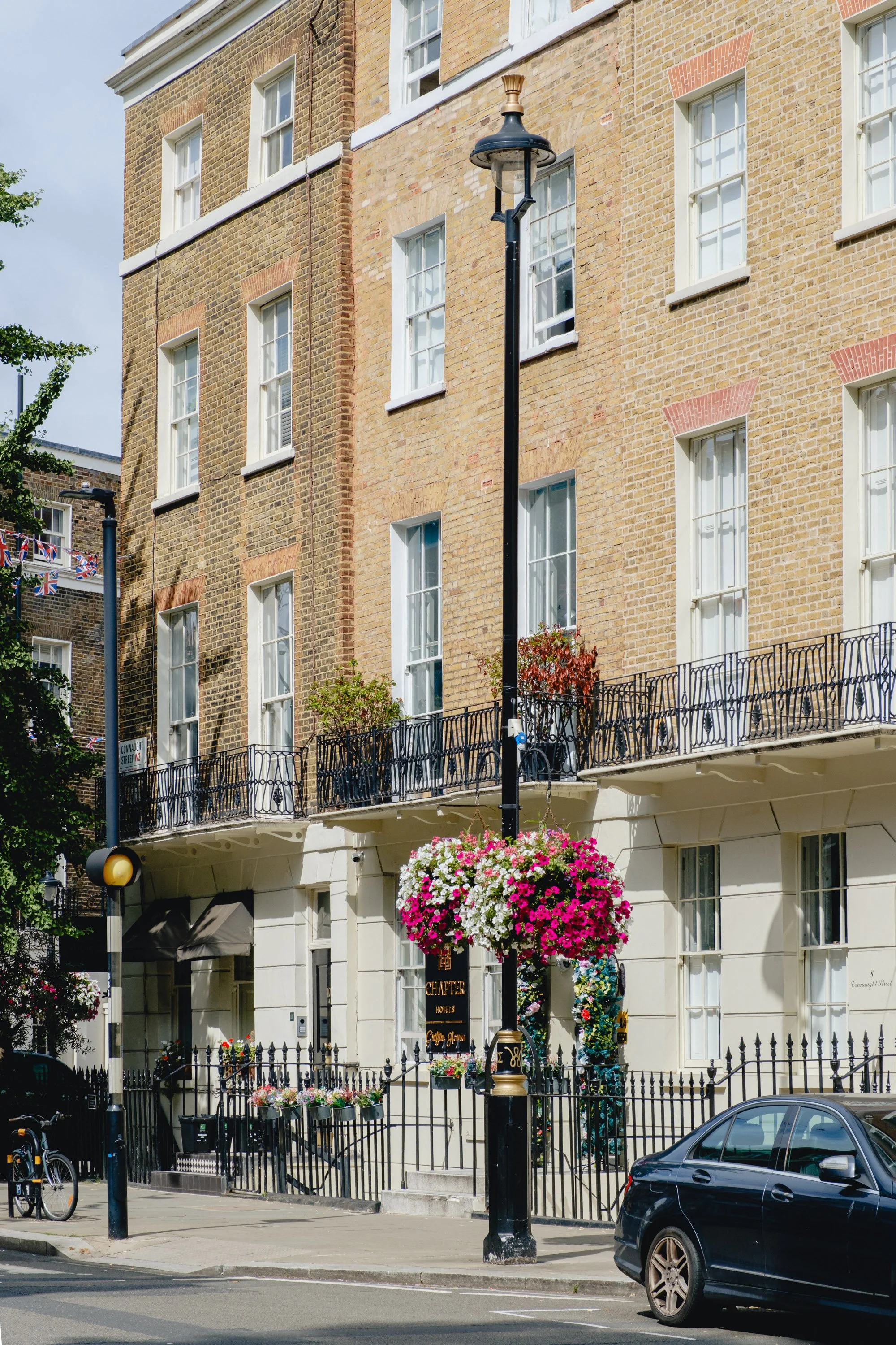 A street corner with a lamp post decorated with hanging flower baskets, a parked car, a black fence surrounding a building with multiple white window frames and a small balcony, and a bike parked on the sidewalk.