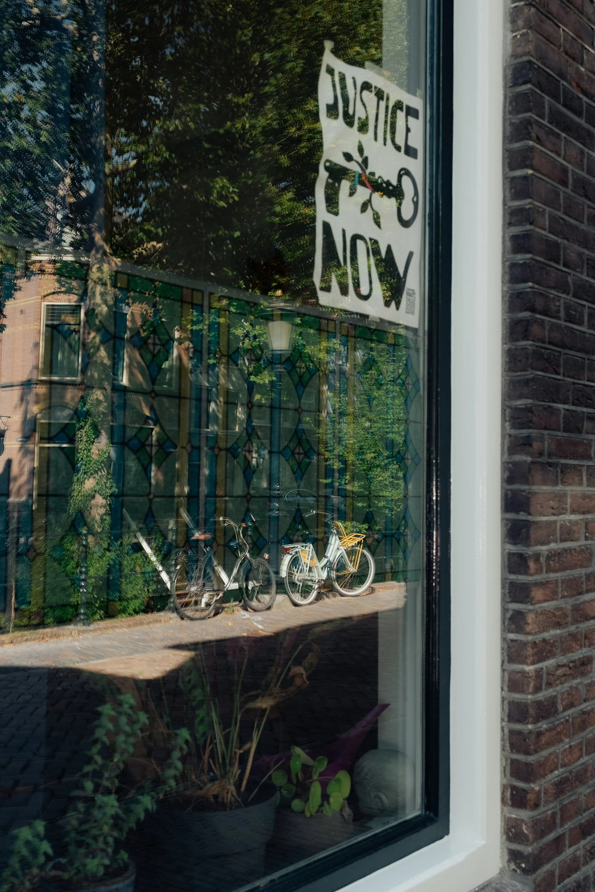 Reflection of three bicycles parked outside, seen through a window with a sign that reads 'Justice Now' in handwritten style.