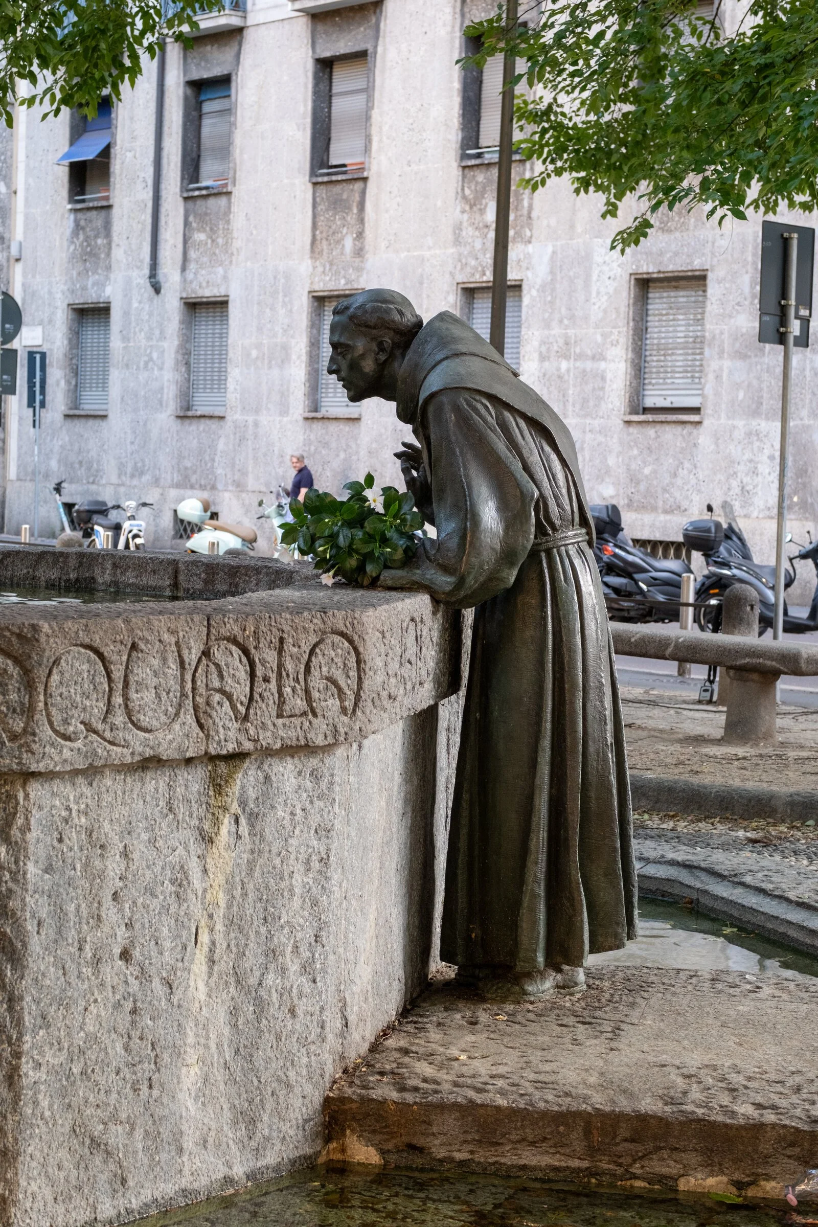 A bronze statue of a hooded friar bends over a stone fountain edge inscribed with lettering, tending a bundle of flowers