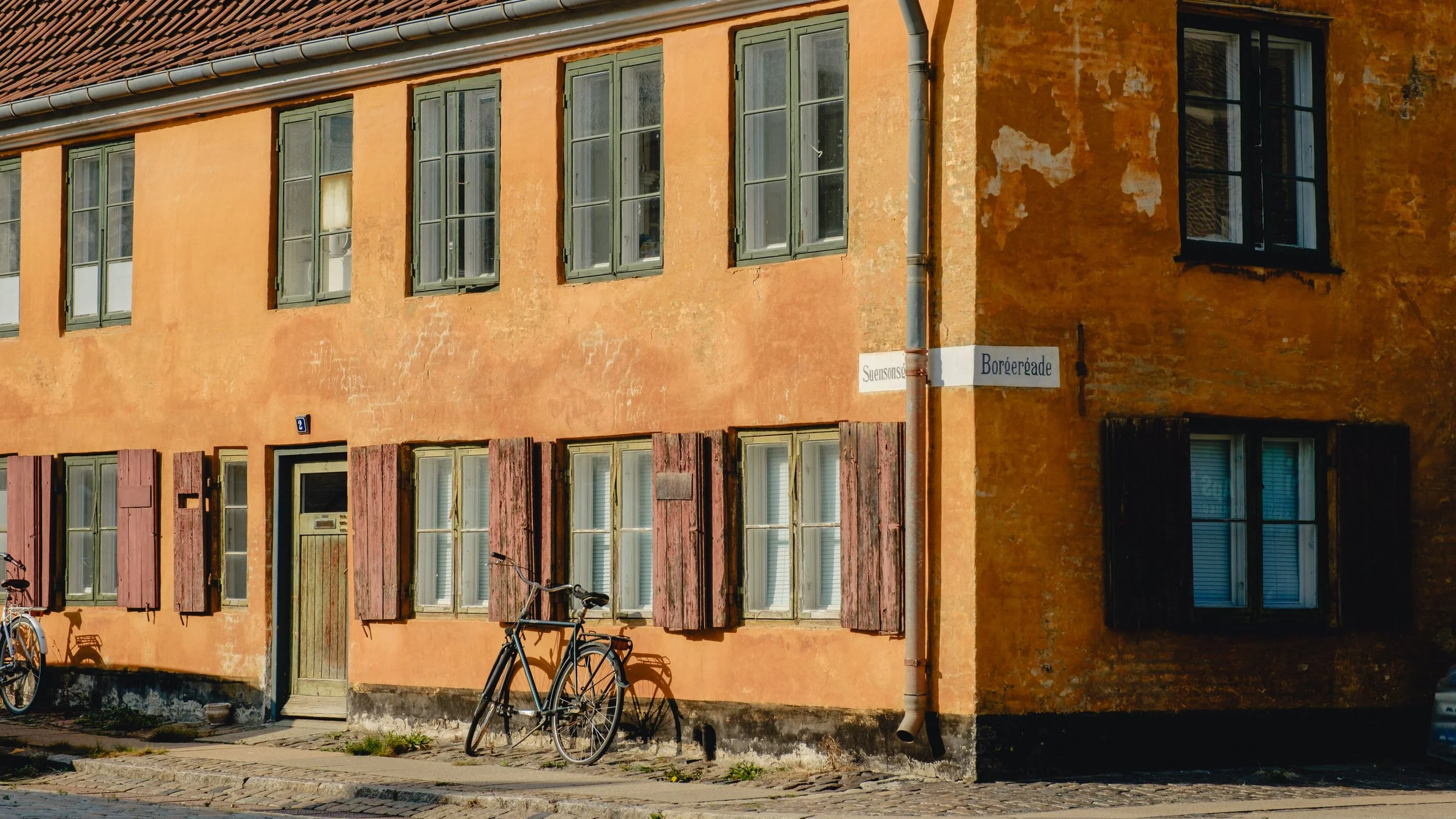 A two-story orange building with multiple green-framed windows, some open with shutters, and bicycles parked outside. There are street signs for Suensonsgade and Børgergade on the corner.