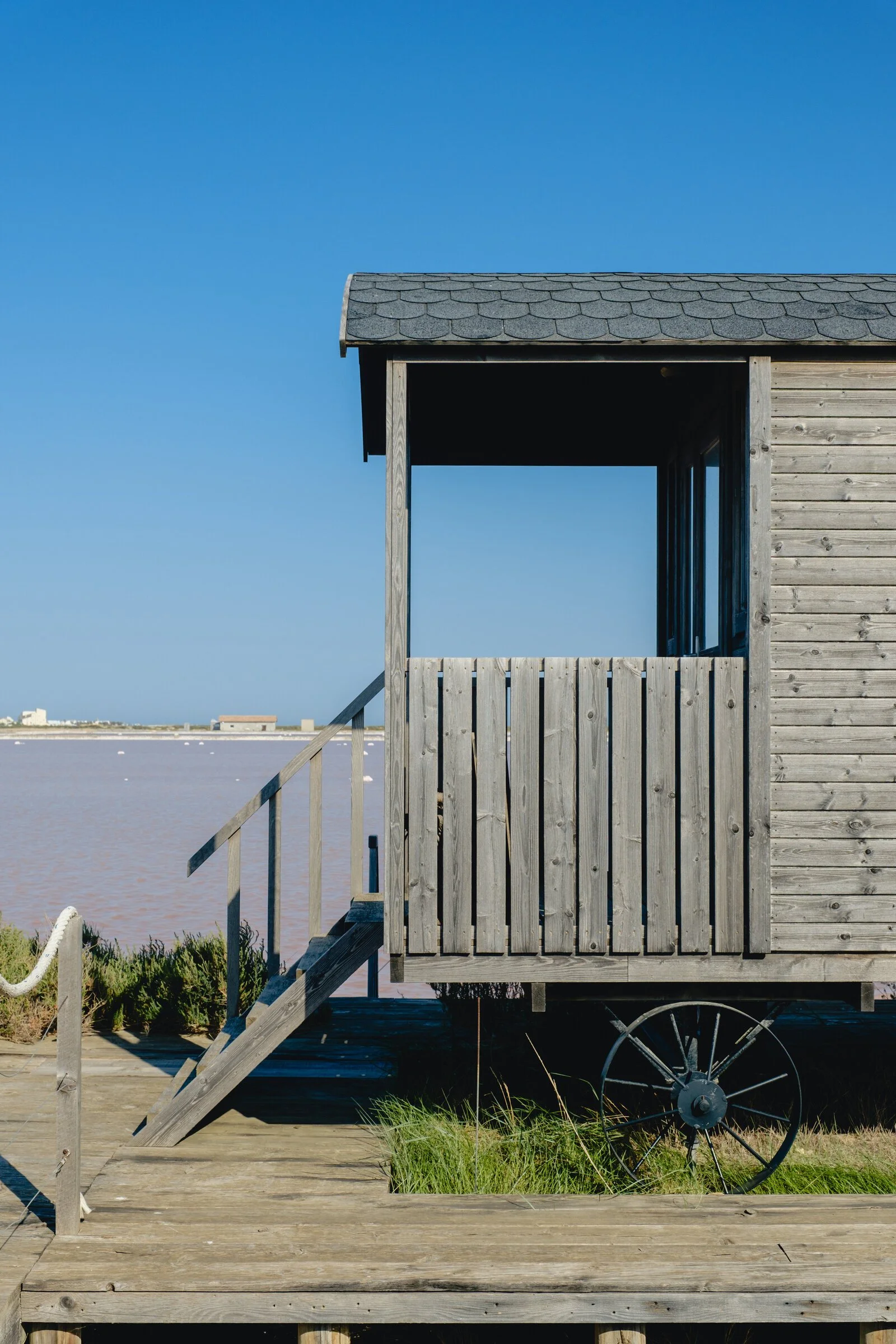 A wooden fisherman's cabin on stilts stands beside pink-tinted salt pan water under a clear blue sky