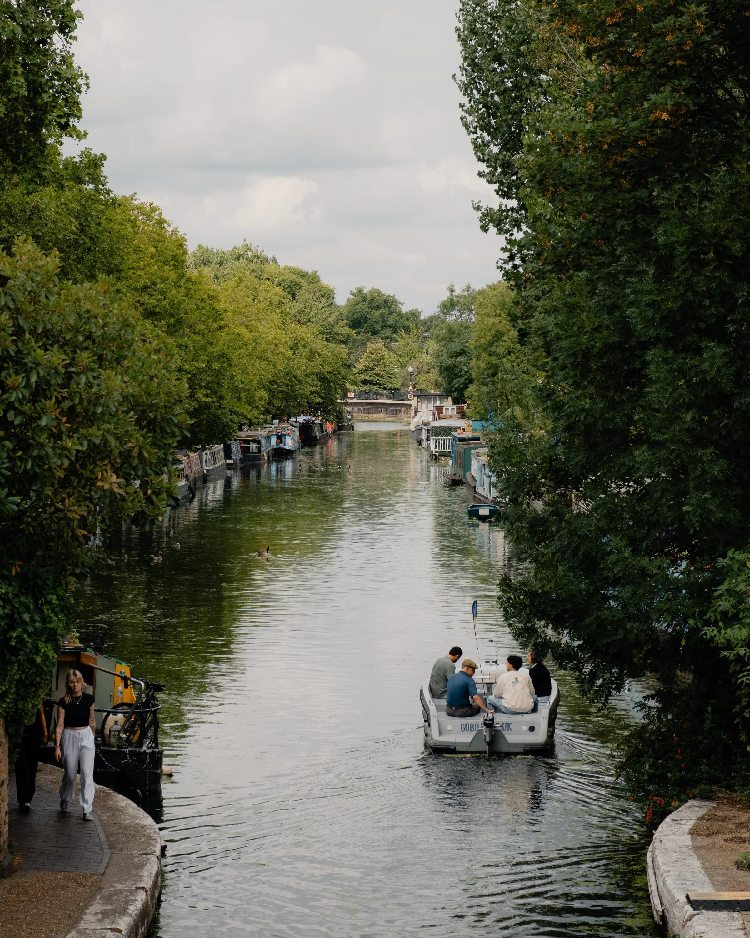 A canal with boats on both sides, trees lining the banks, and people enjoying a boat ride on a small motorboat in the foreground.