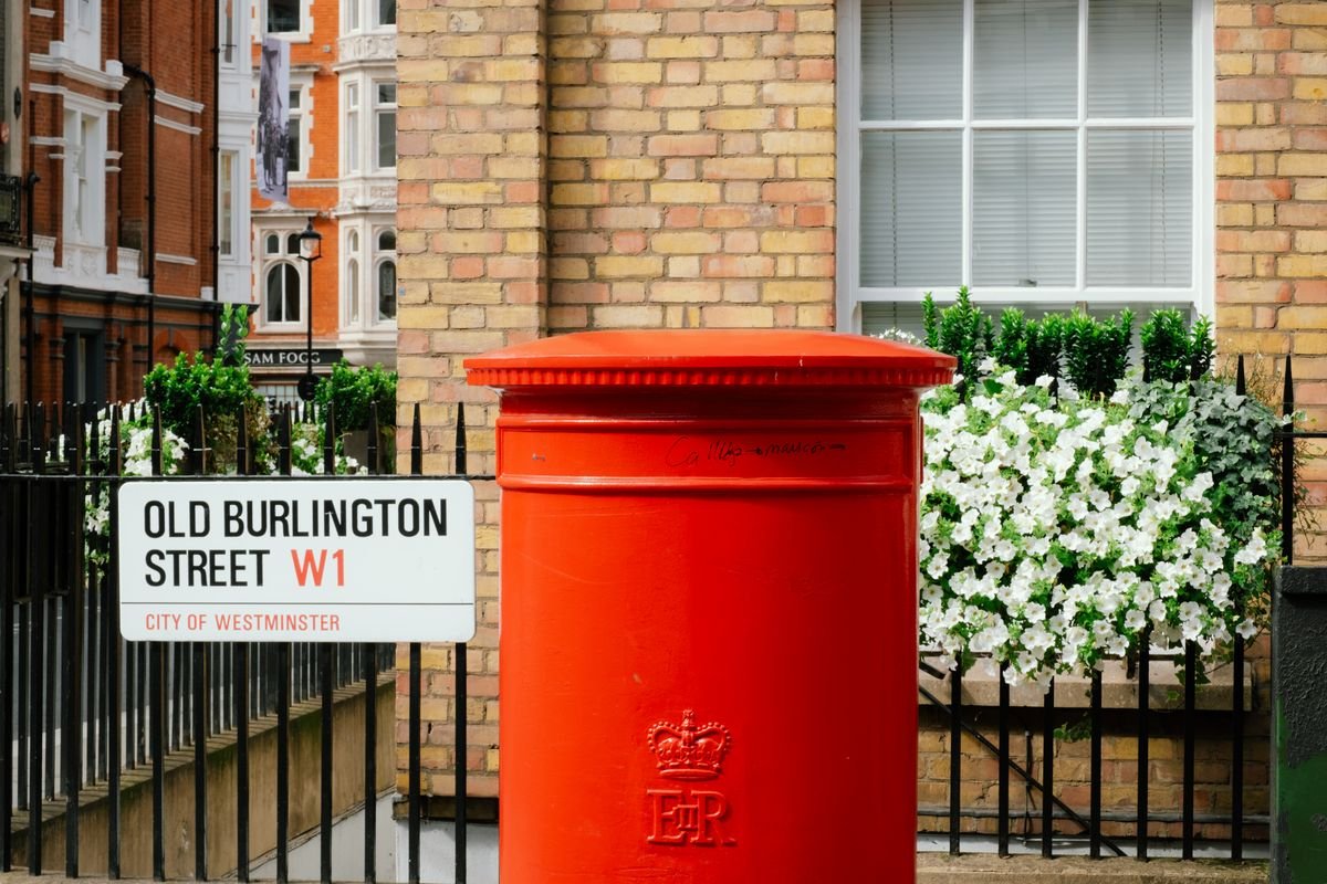 A red pillar postbox stands before an Old Burlington Street sign, white flowers behind