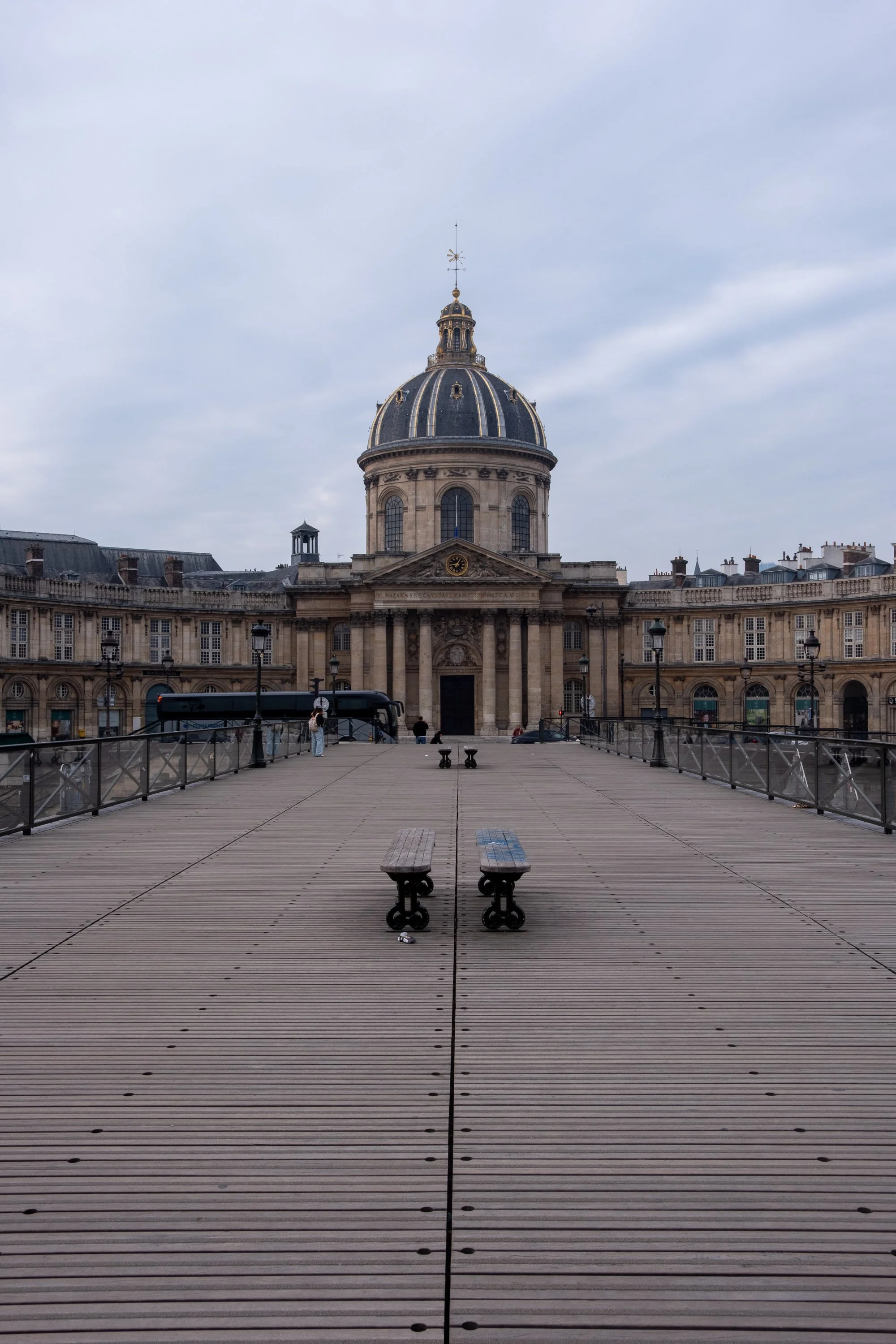 View of a historic building with a large domed roof, seen from a wooden walkway flanked by benches and lampposts.