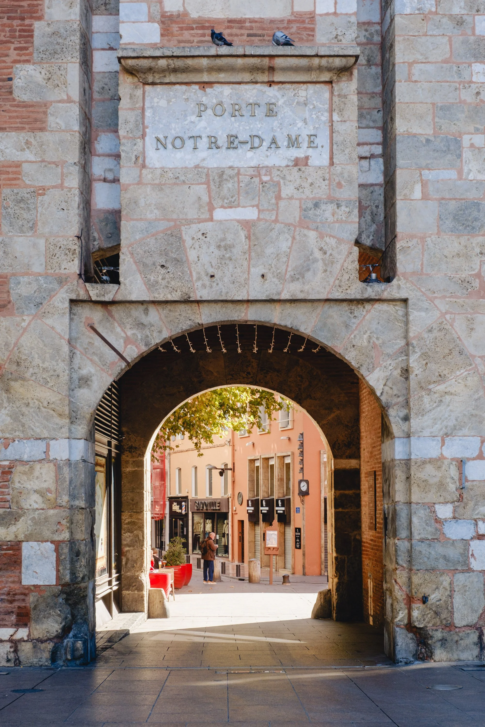 Arched stone gateway with the inscription 'Porte Notre-Dame,' leading to a street scene with colorful buildings, a person walking, trees, and outdoor seating.