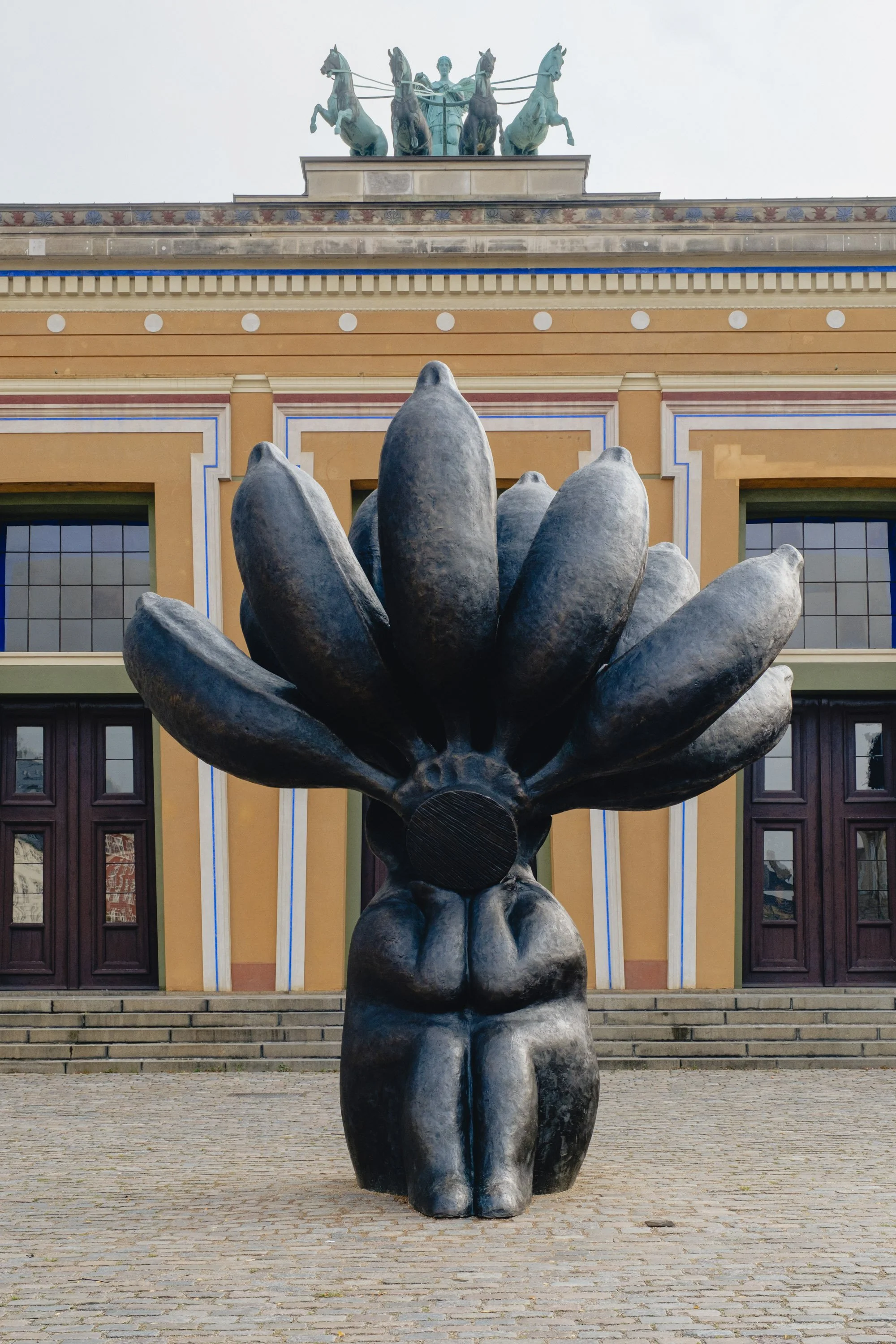 A large black sculpture of a woman with an abstract flower-shaped head standing on a cobblestone ground in front of a colorful building with large windows and doors. A bronze chariot sculpture with horses and a human figure is atop the building.