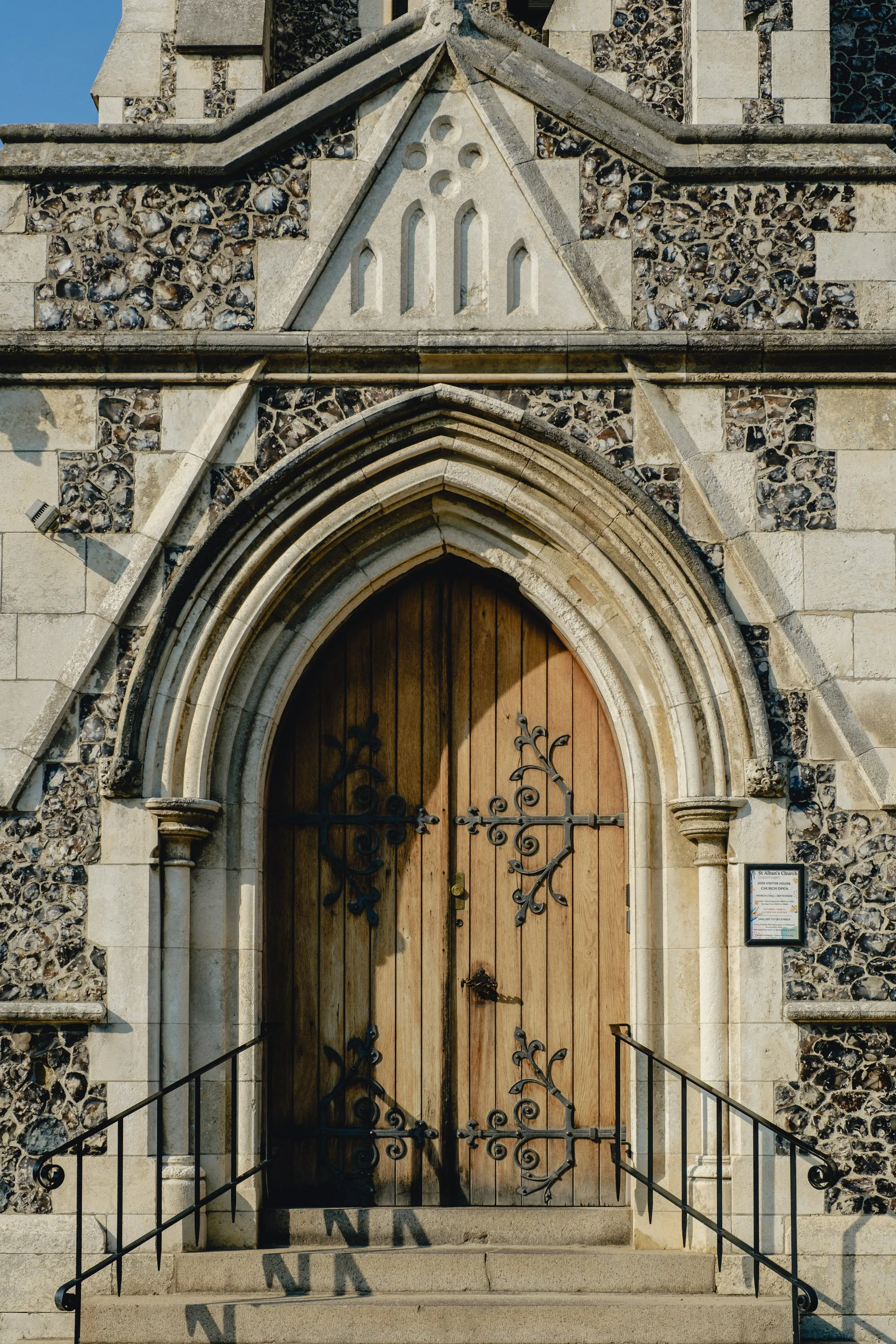 Front view of a stone church entrance with wooden arched door and decorative black metalwork, surrounded by stone walls and steps.