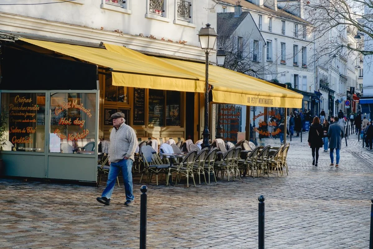 A man walks past a Montmartre café with a yellow awning and wicker bistro chairs