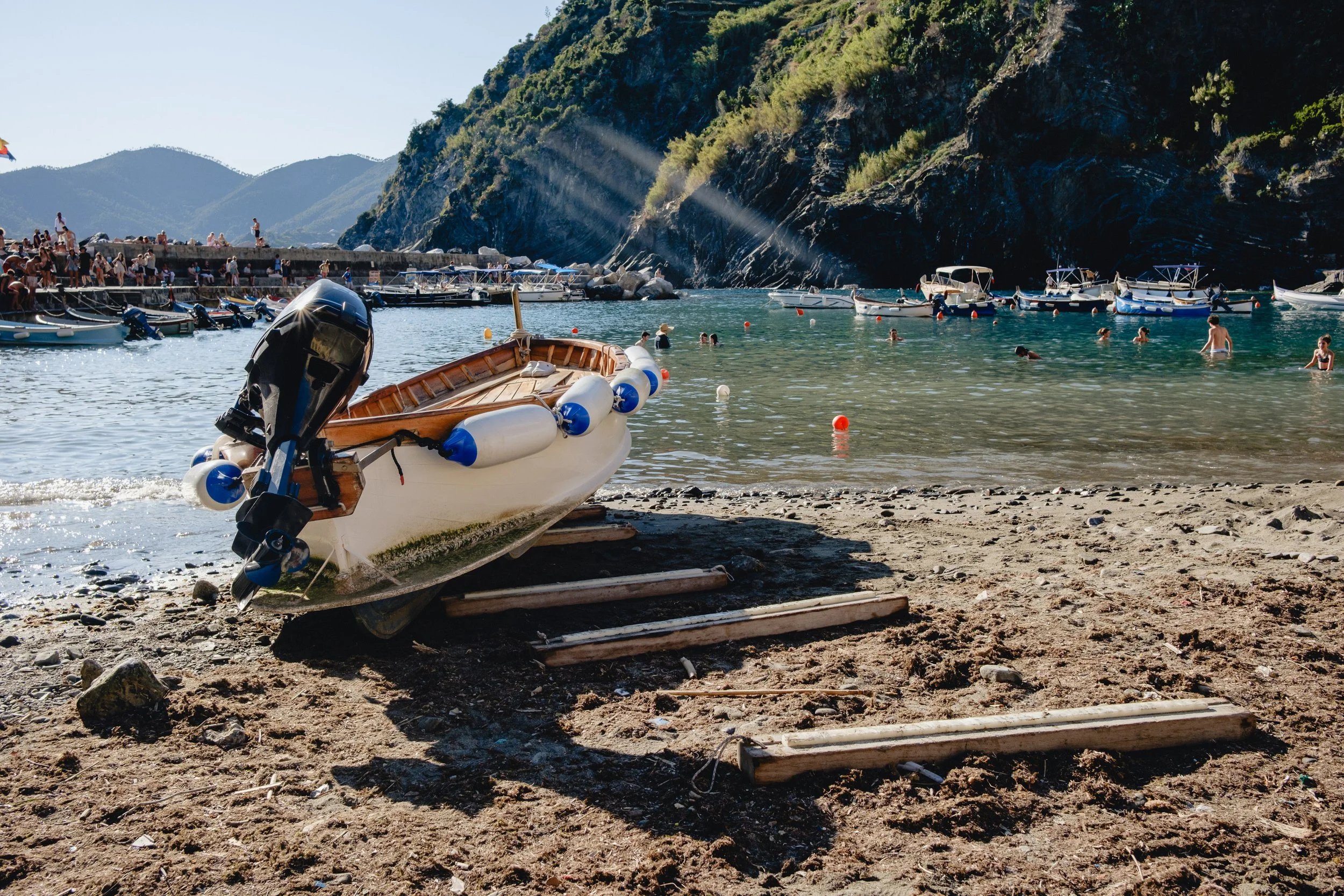 A small boat with an outboard motor resting on the sandy beach near the water, with several boats anchored in a bay surrounded by green hills and many people swimming and relaxing in the water.