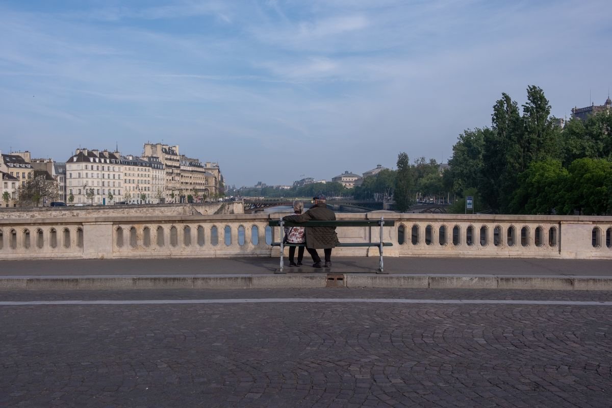 An elderly couple sit together on a bench overlooking the Seine from Pont Neuf