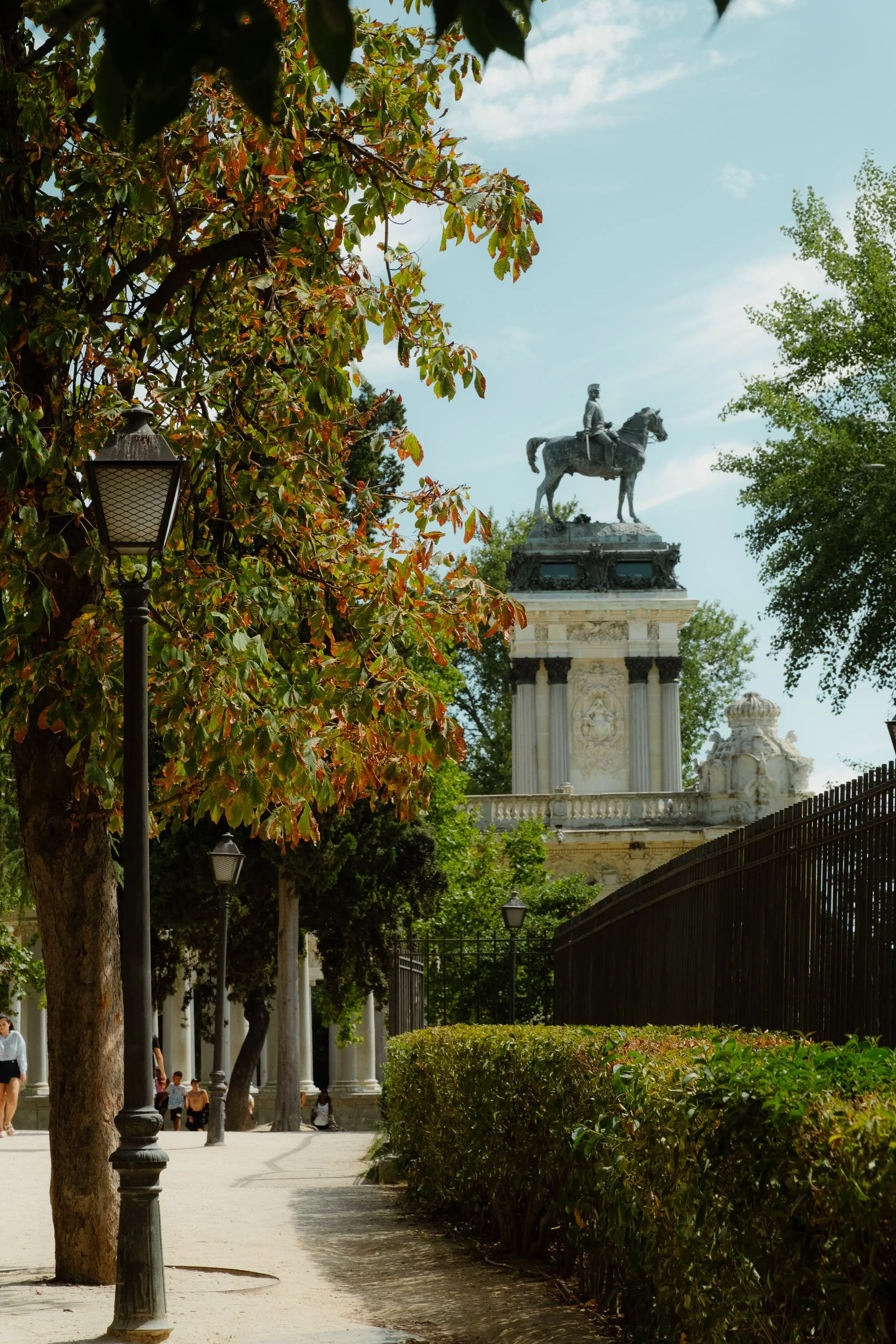 A park with trees, street lamps, and a pathway, leading to a monument with an equestrian statue on top, featuring a person riding a horse.