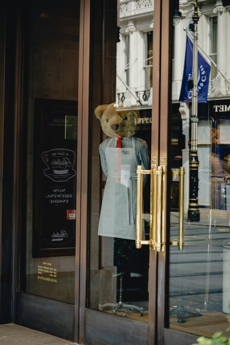 A bow-tied teddy bear mannequin stands behind a shopfront's glass doors
