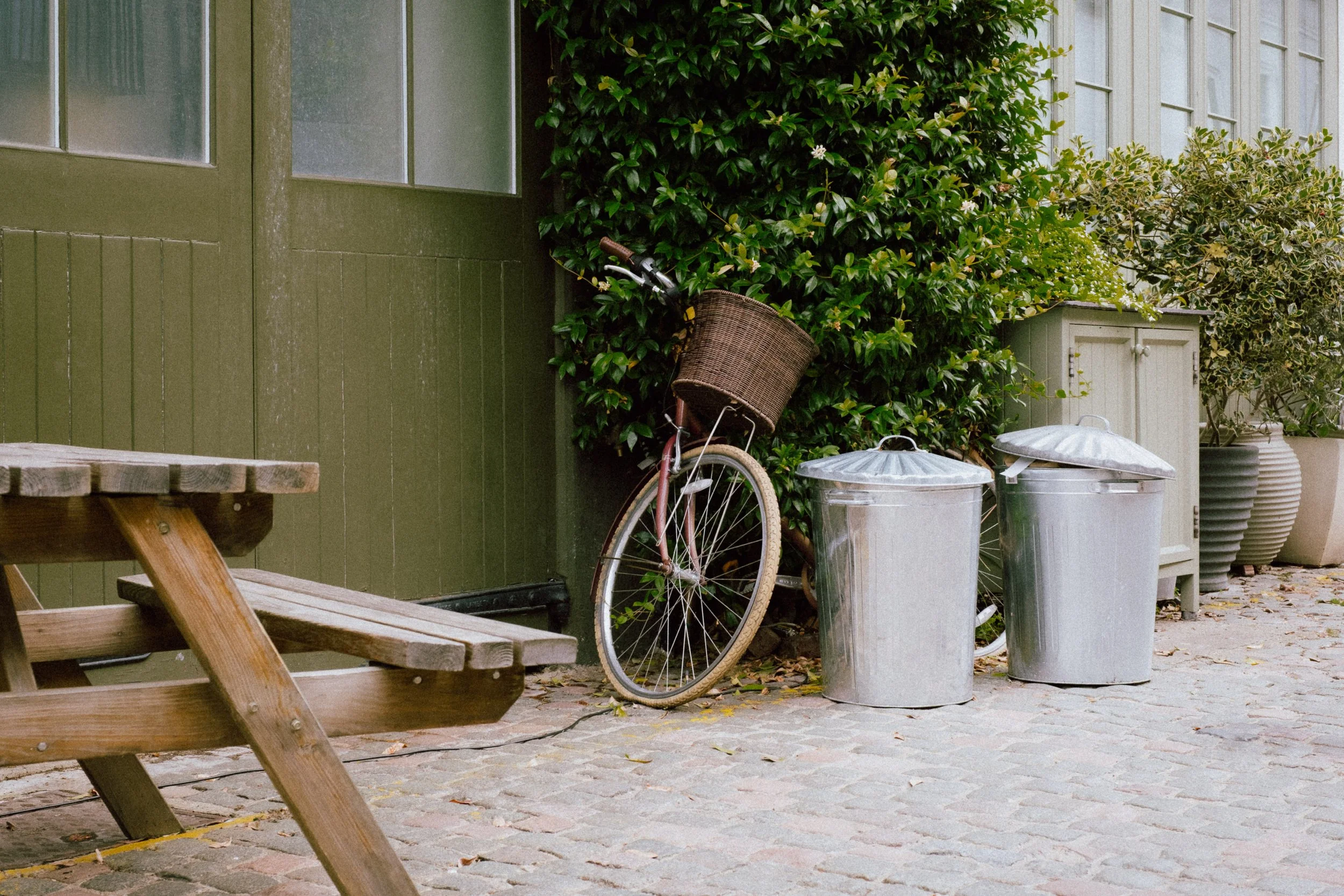 An outdoor scene with a transparent bicycle with a wicker basket leaning against a green house wall, two metal trash cans with lids, a wooden picnic table and benches, green bushes, and large potted plants.