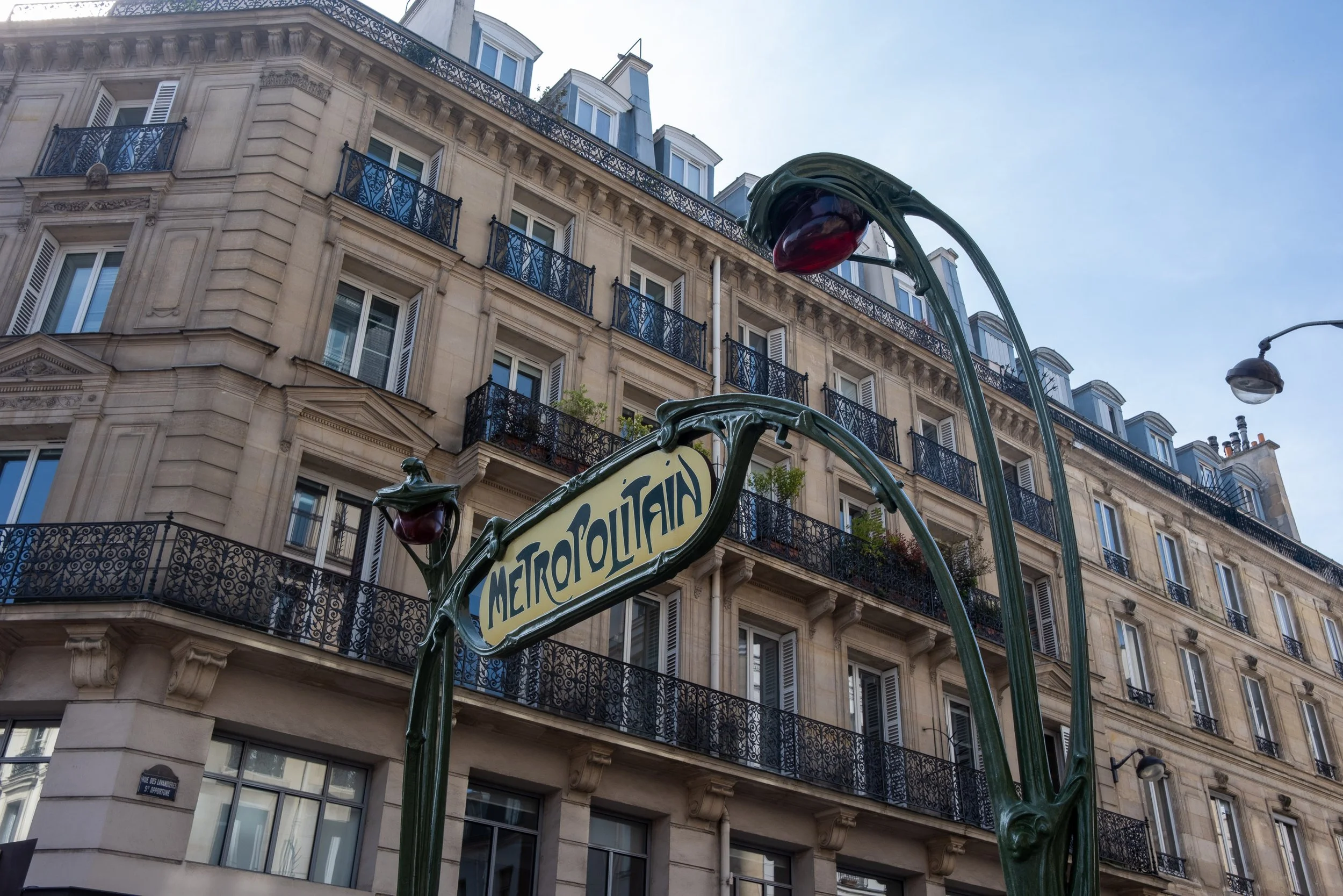 Paris Métro sign with artistic floral design, ornate building with balconies and window shutters in the background, street lamp, clear sky.
