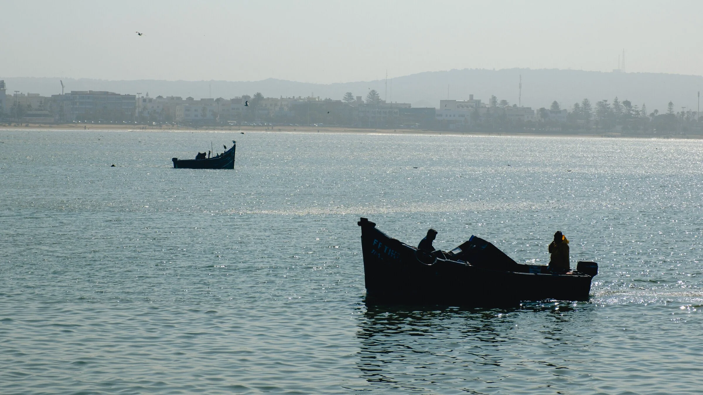 Small fishing boats sit silhouetted on shimmering, backlit harbor water