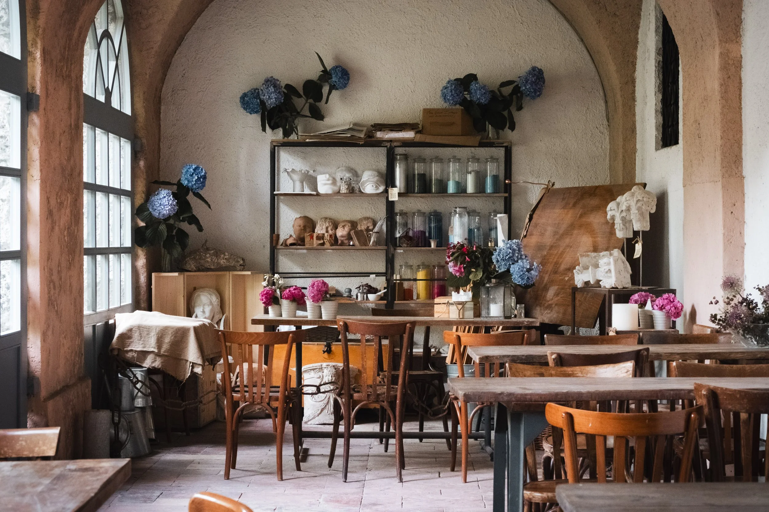 Interior of a cozy cafe or boutique with wooden tables and chairs, decorated with blue and pink hydrangea floral arrangements, shelves with artistic and vintage items, bust sculptures, and large windows letting in natural light.