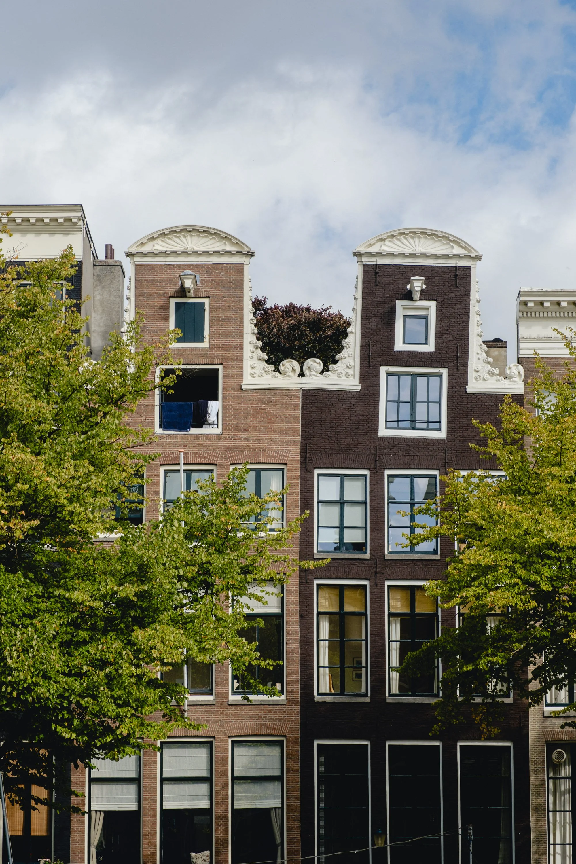 Tall historic brick apartment building with large windows and decorative white architectural details, surrounded by green trees, under a cloudy sky.