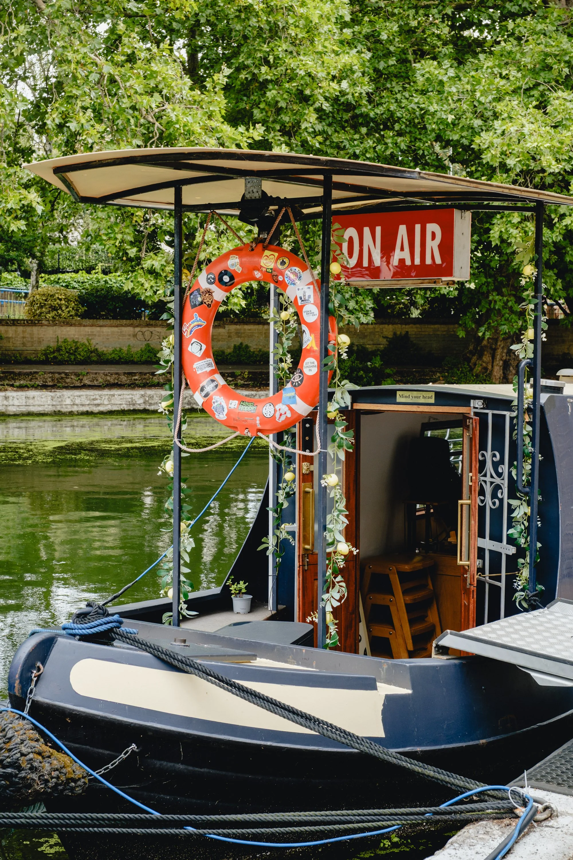 A boat decorated with a life ring covered in stickers, an 'On Air' sign, and surrounded by greenery, docked on a river.