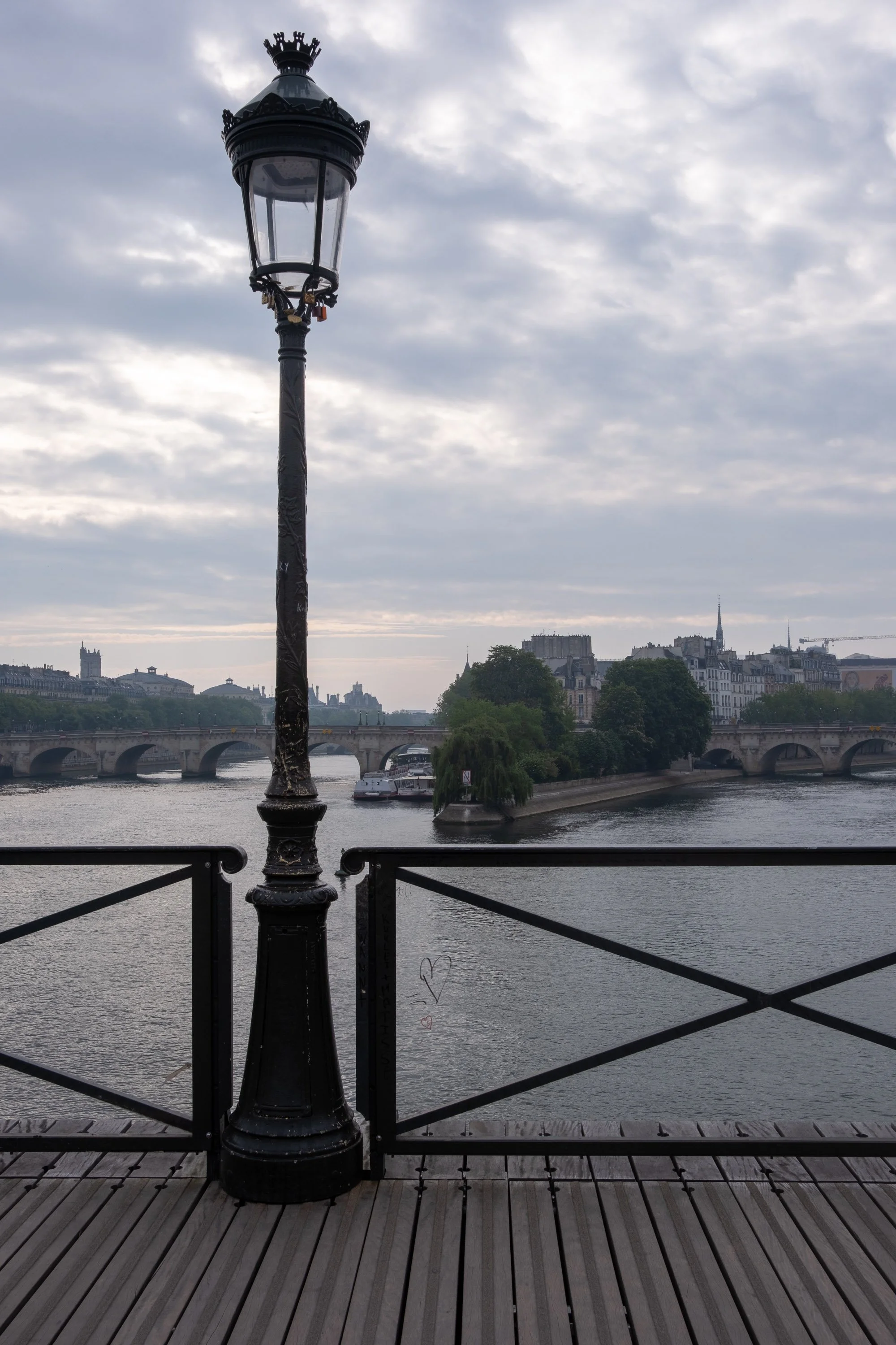 A black street lamp on a wooden deck beside a river, with a bridge, boats, and buildings in the background on a cloudy day.