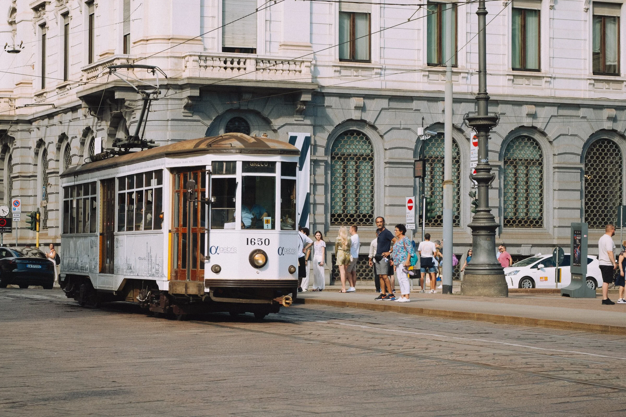 A vintage orange-and-cream Milan tram crosses a cobbled piazza as pedestrians wait to cross
