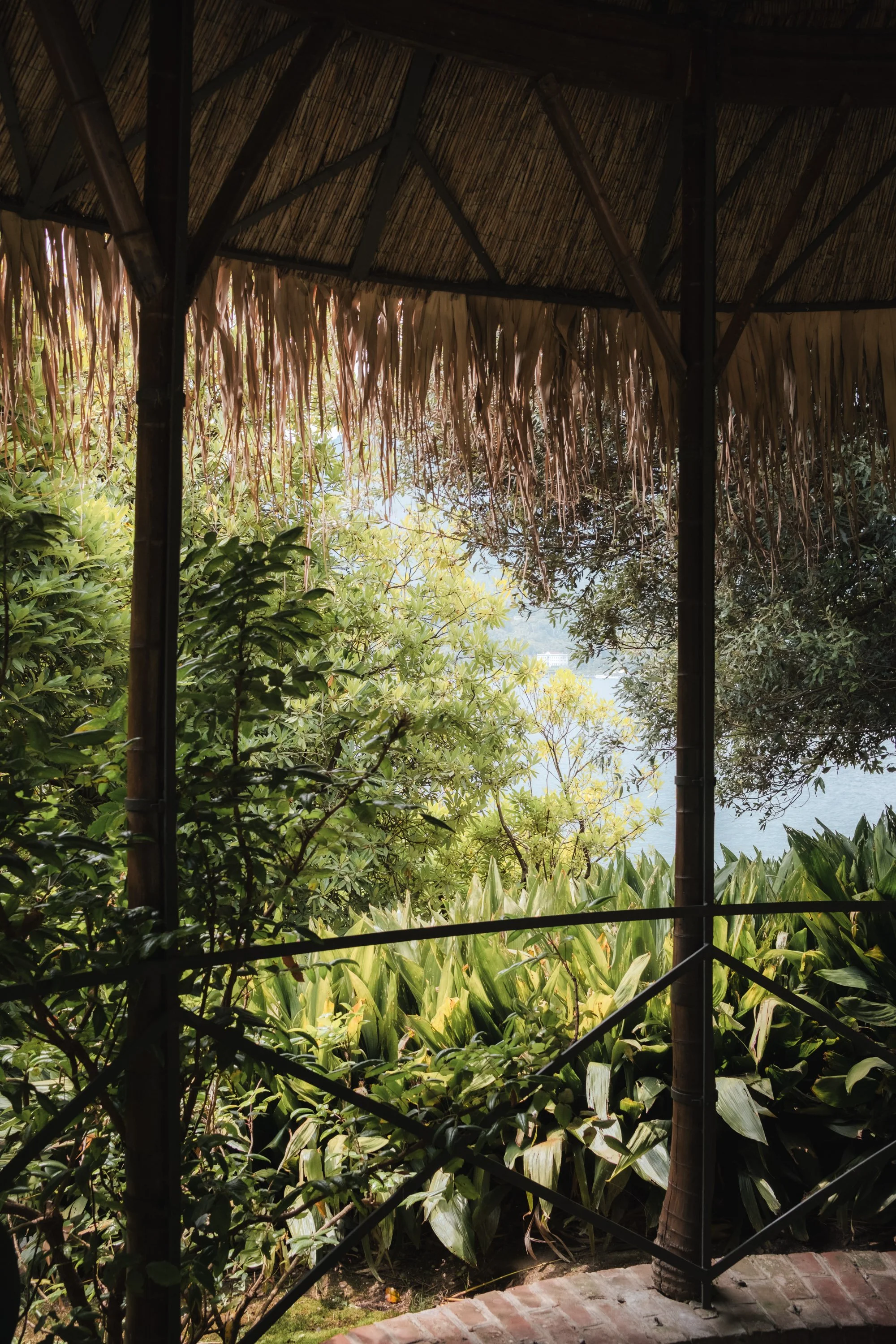 View from a bamboo and thatched-roof hut overlooking lush green foliage and a body of water in the distance.