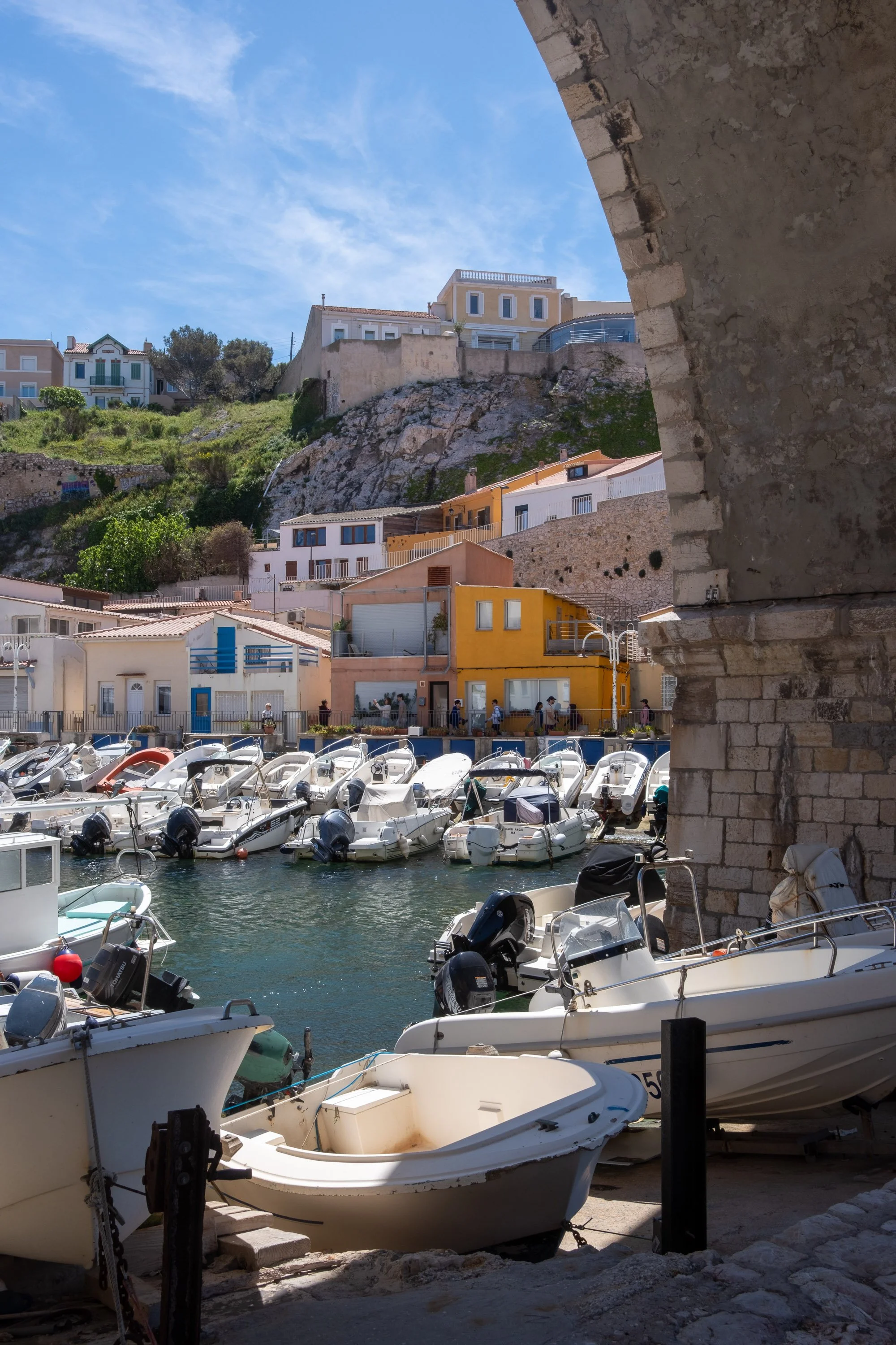 Boats docked in a harbor with colorful buildings on a hillside in the background under a blue sky.